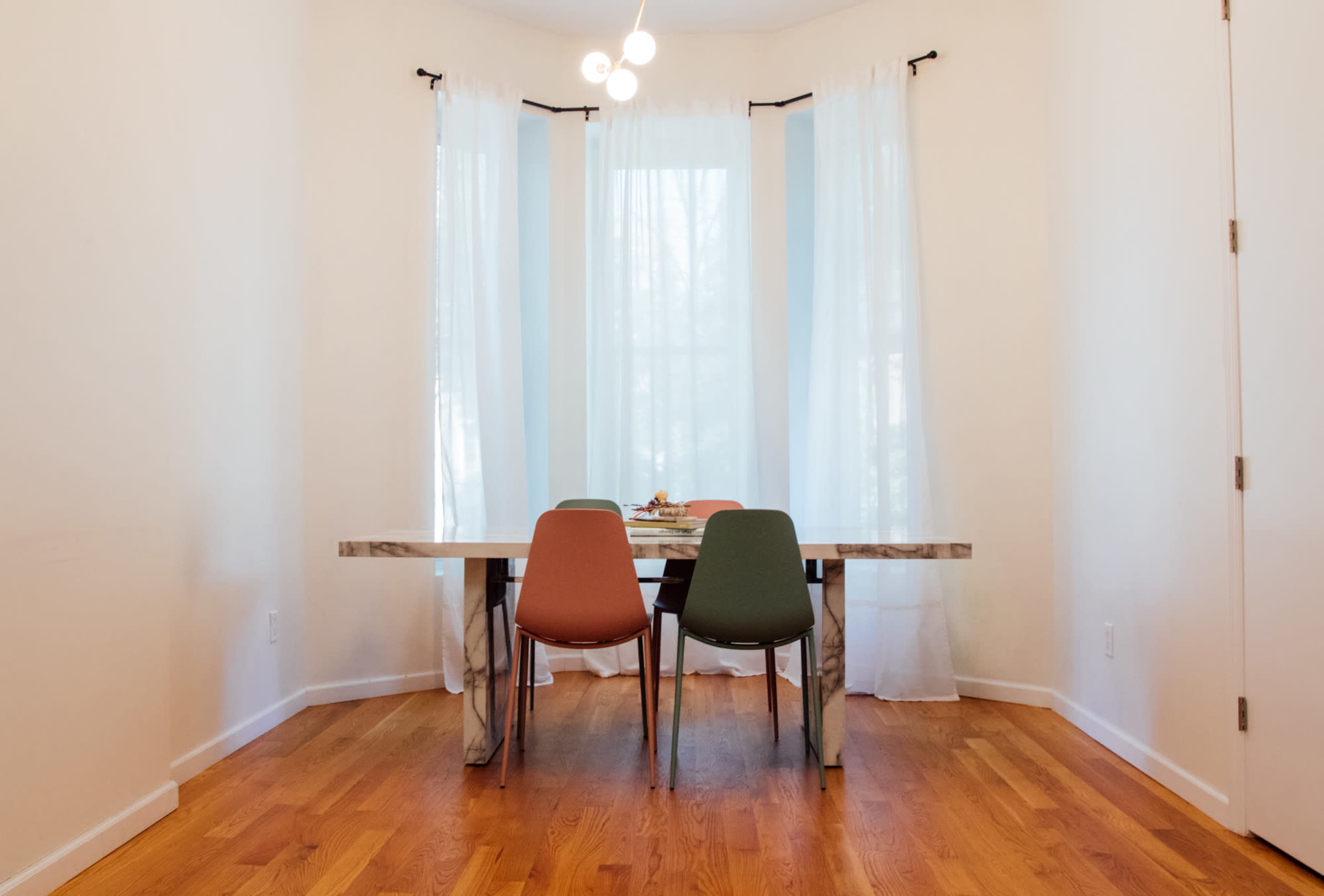 A dining area features a table with two chairs, large windows covered by sheer curtains, and hardwood flooring.