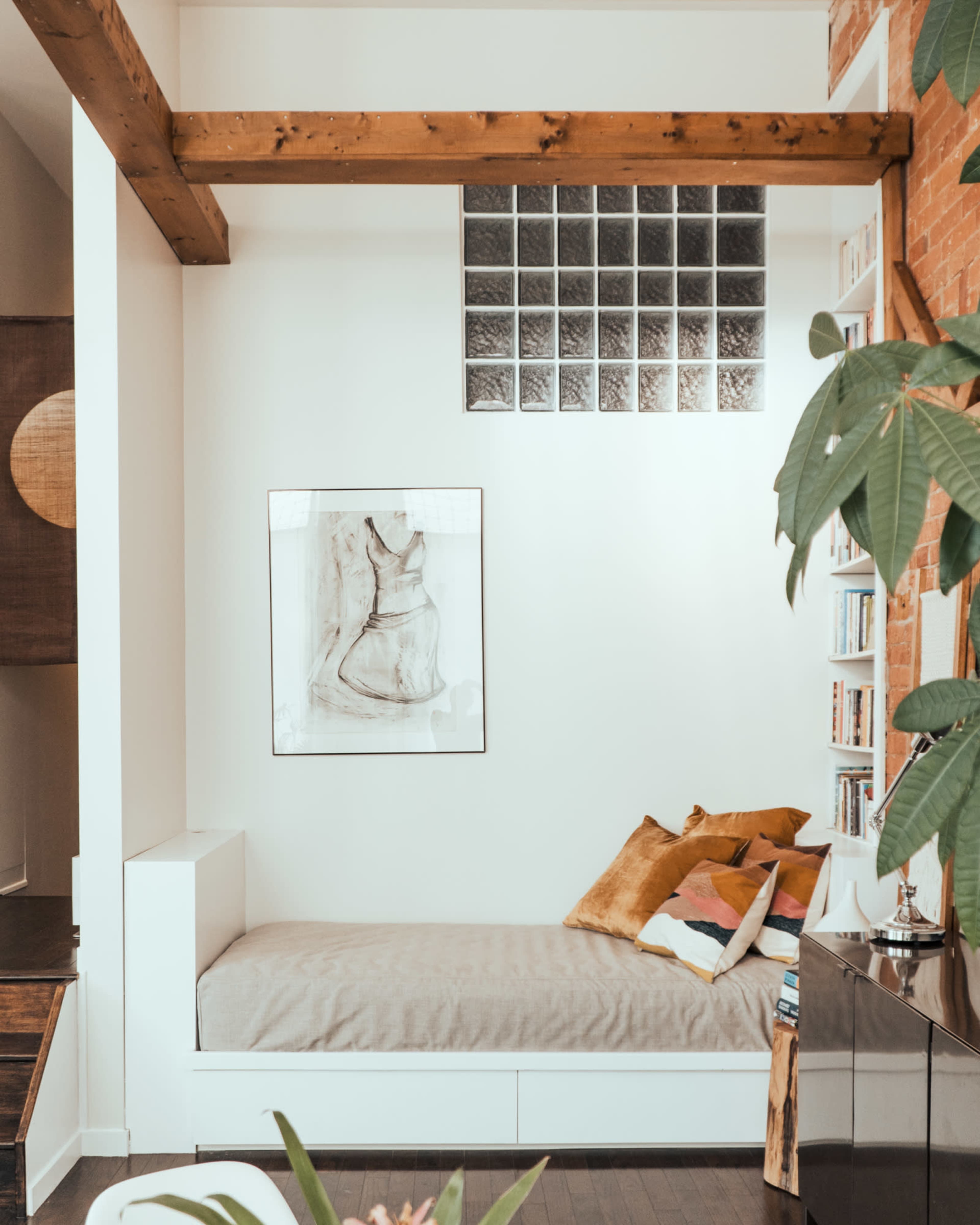 A cozy nook with a daybed adorned with pillows, positioned near a wall featuring a glass block window and a framed artwork above.