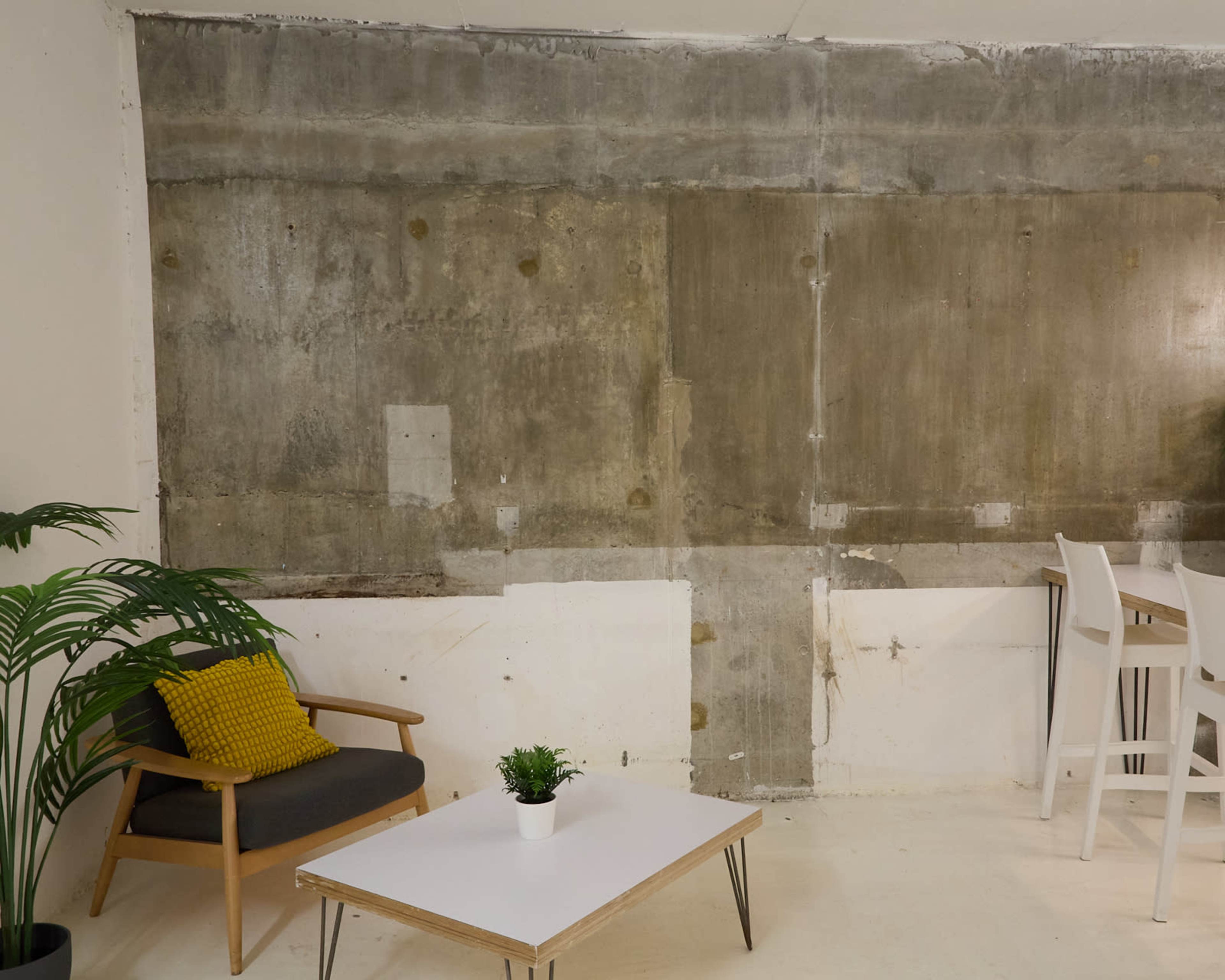 The image depicts a minimalist interior with a concrete wall, a wooden table, a small plant, and a chair next to a set of white stools.