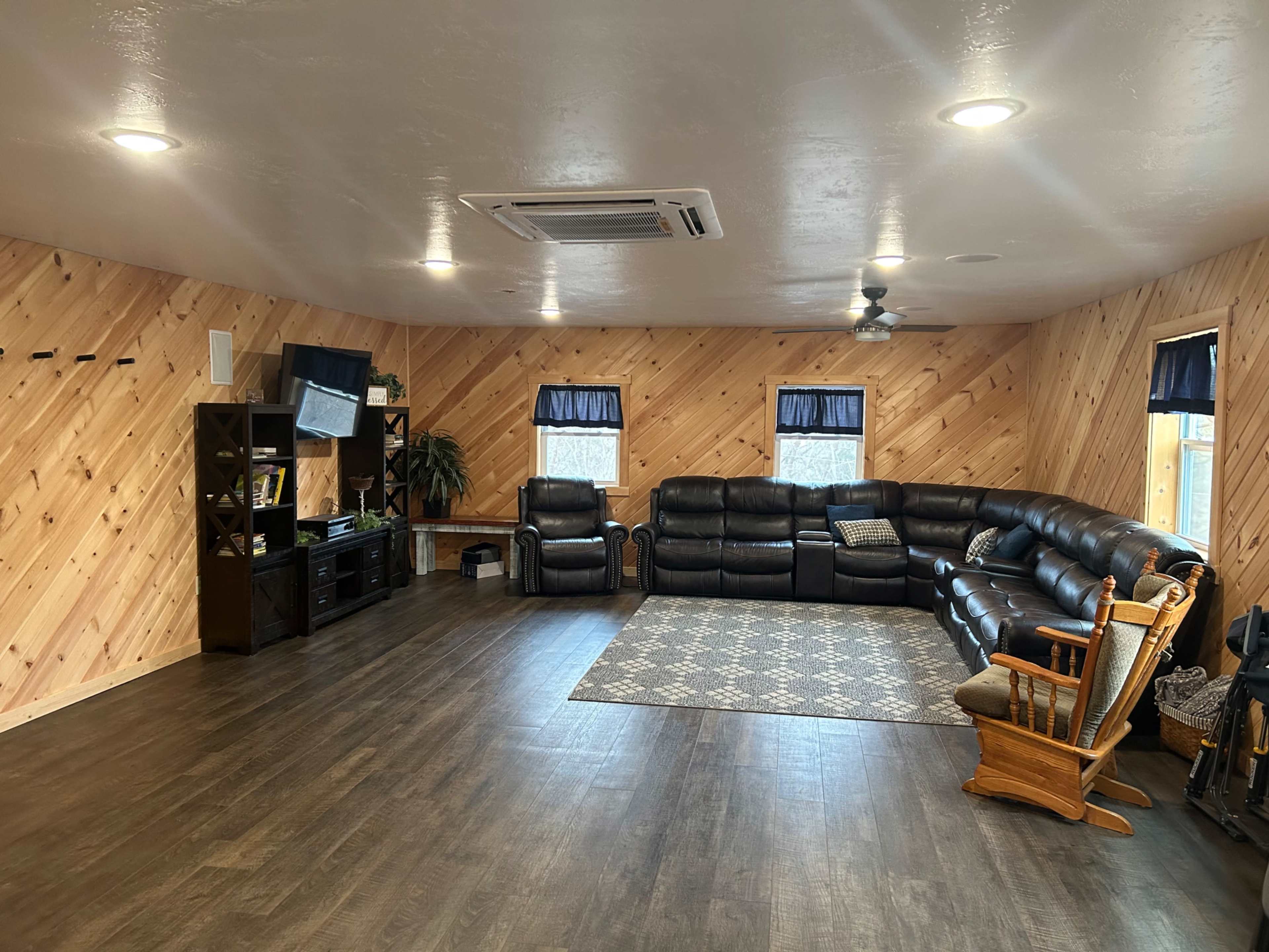 The room features a spacious seating area with a sectional couch, two recliners, a television, and wooden paneling on the walls.