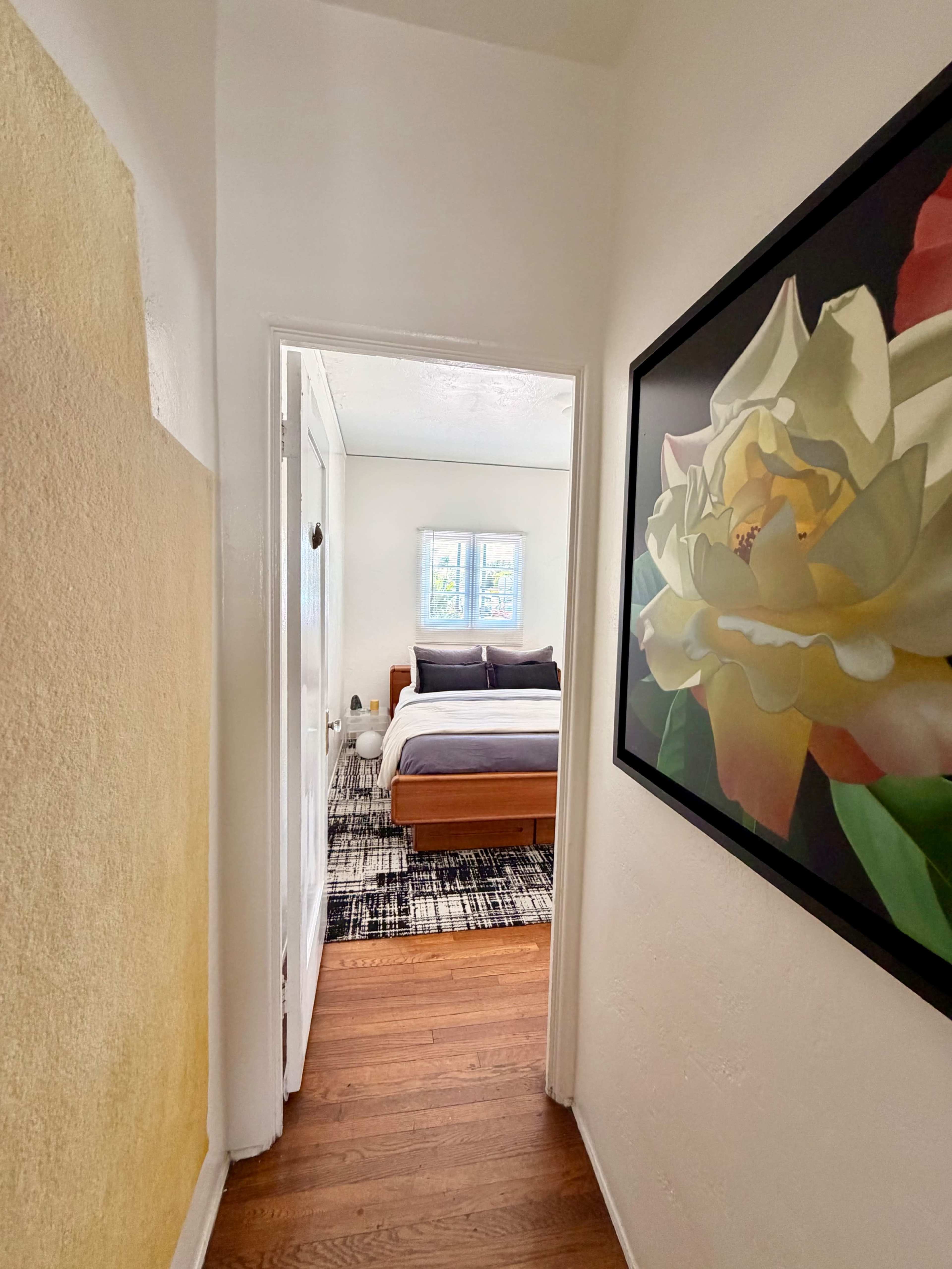 The image shows a narrow hallway leading to a bedroom with a large flower painting on the wall.