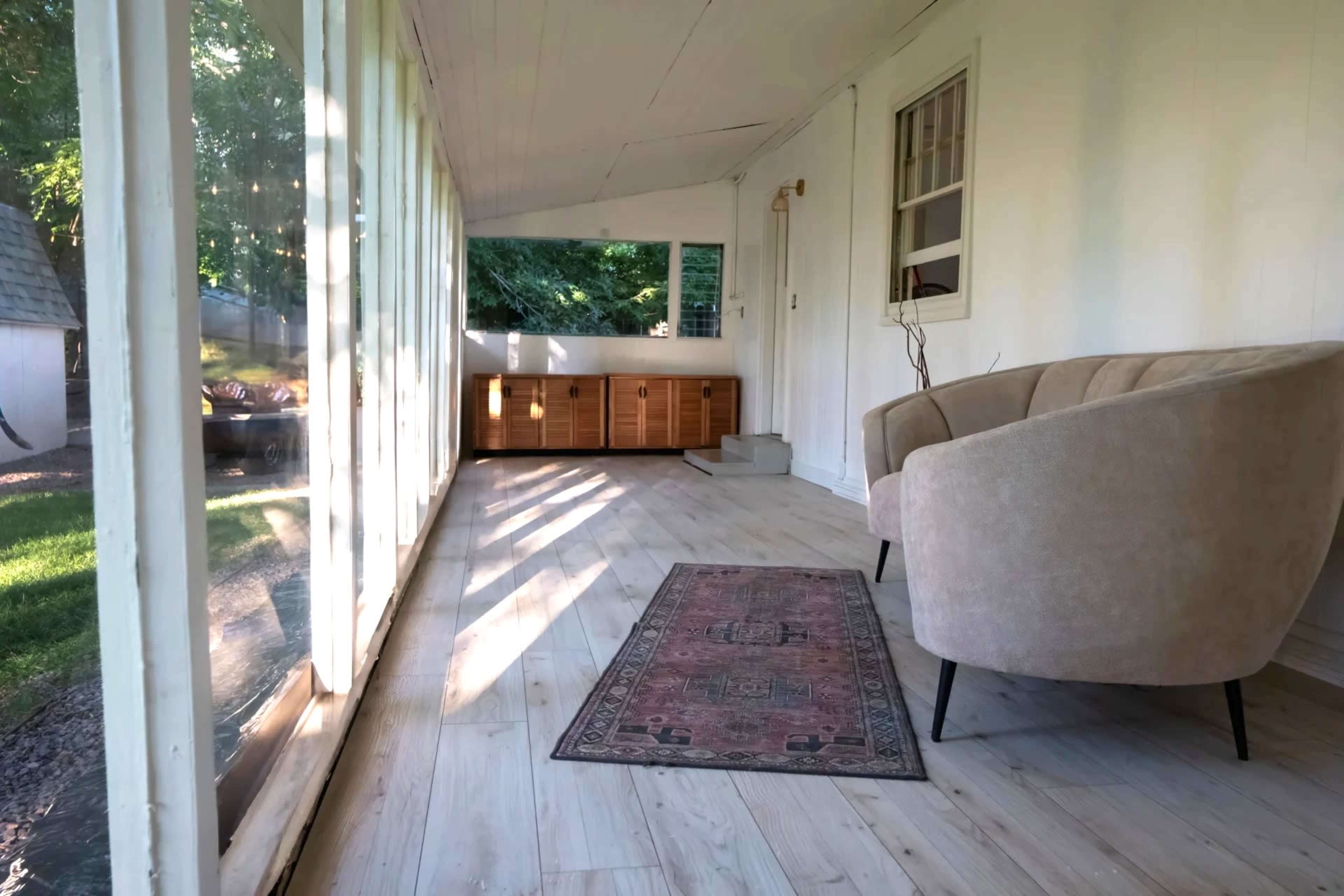 The image shows a sunlit porch with large glass windows, a beige chair, and a patterned rug on a wooden floor.