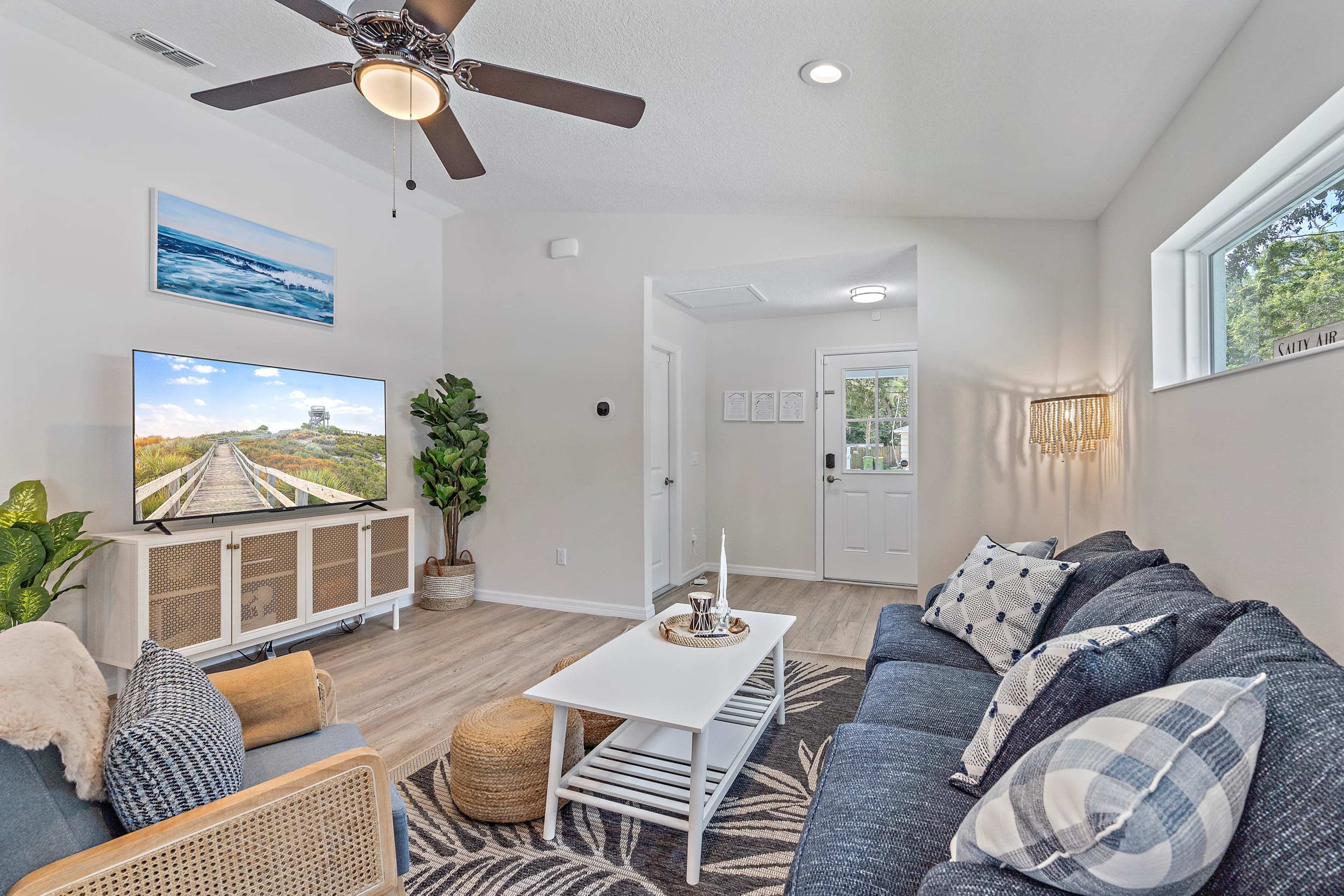 A bright living room with a ceiling fan, a large television, a coffee table, and comfortable seating, along with a doorway leading outside.