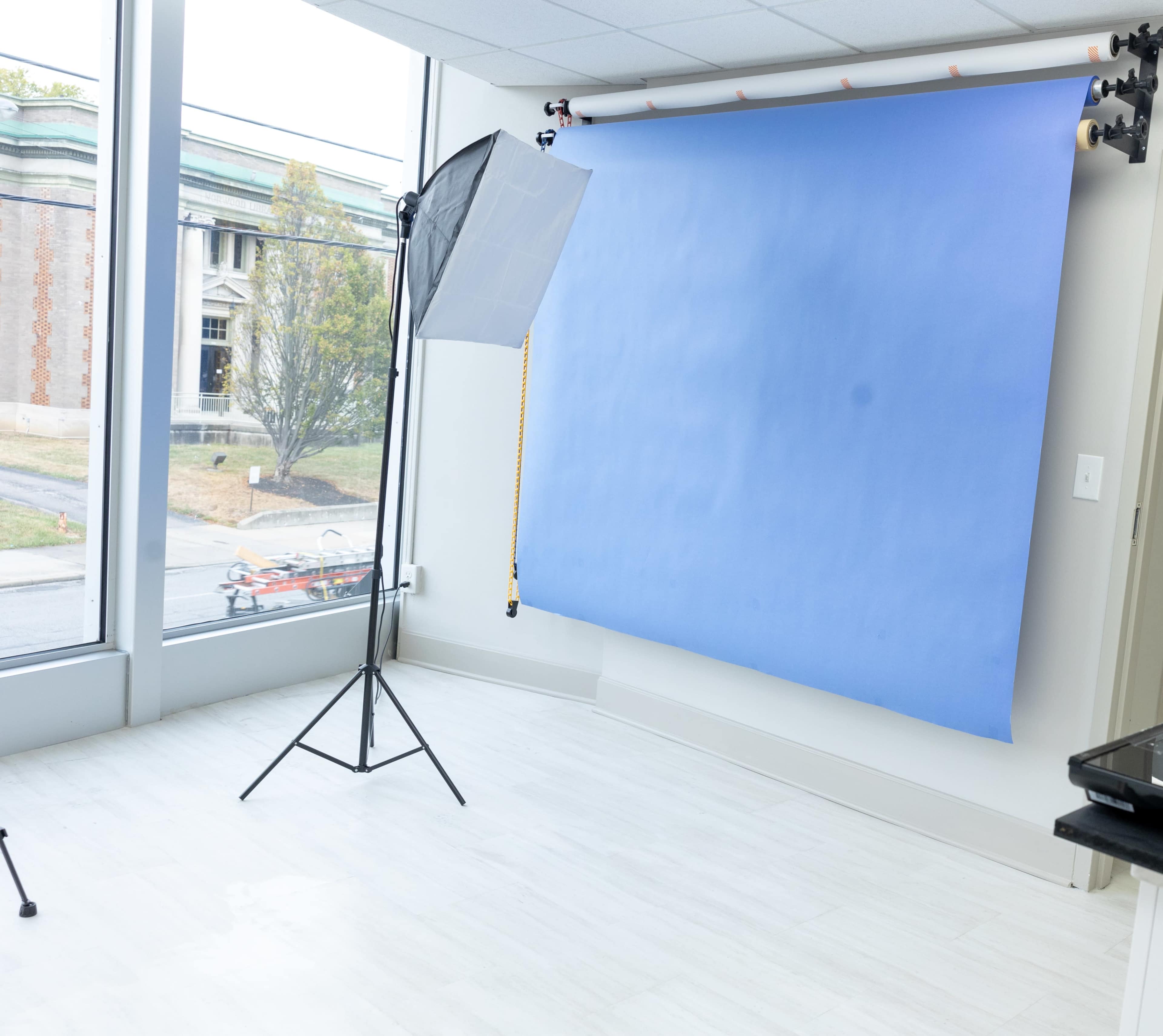 The image shows a bright indoor photography studio setup featuring a blue backdrop, a light stand, and large windows overlooking a street.