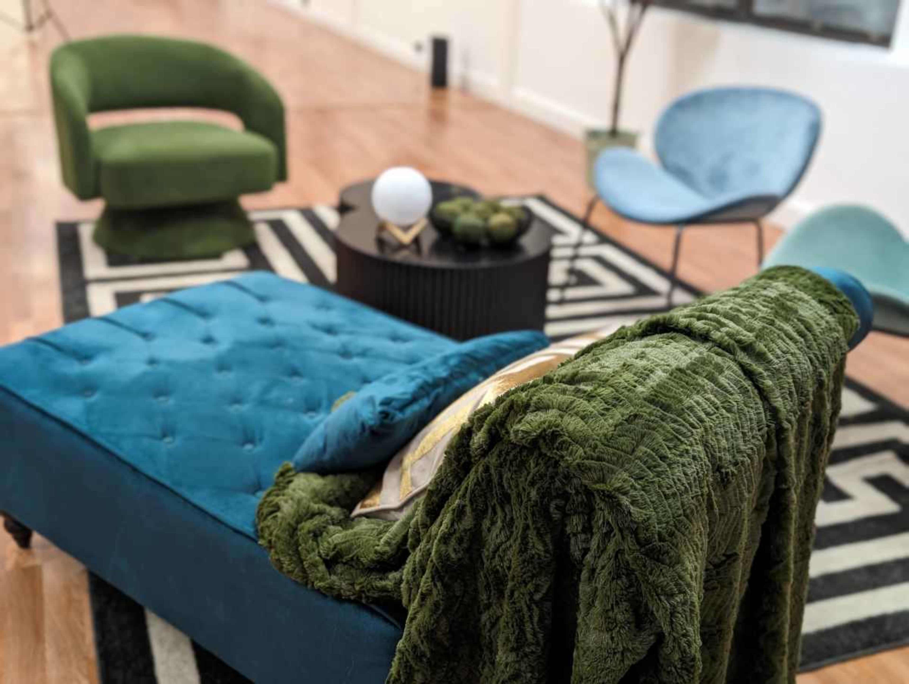 A modern living room setup featuring a teal tufted chaise lounge, a green blanket draped over it, a round black coffee table, and two accent chairs in green and blue, all situated on a patterned rug.