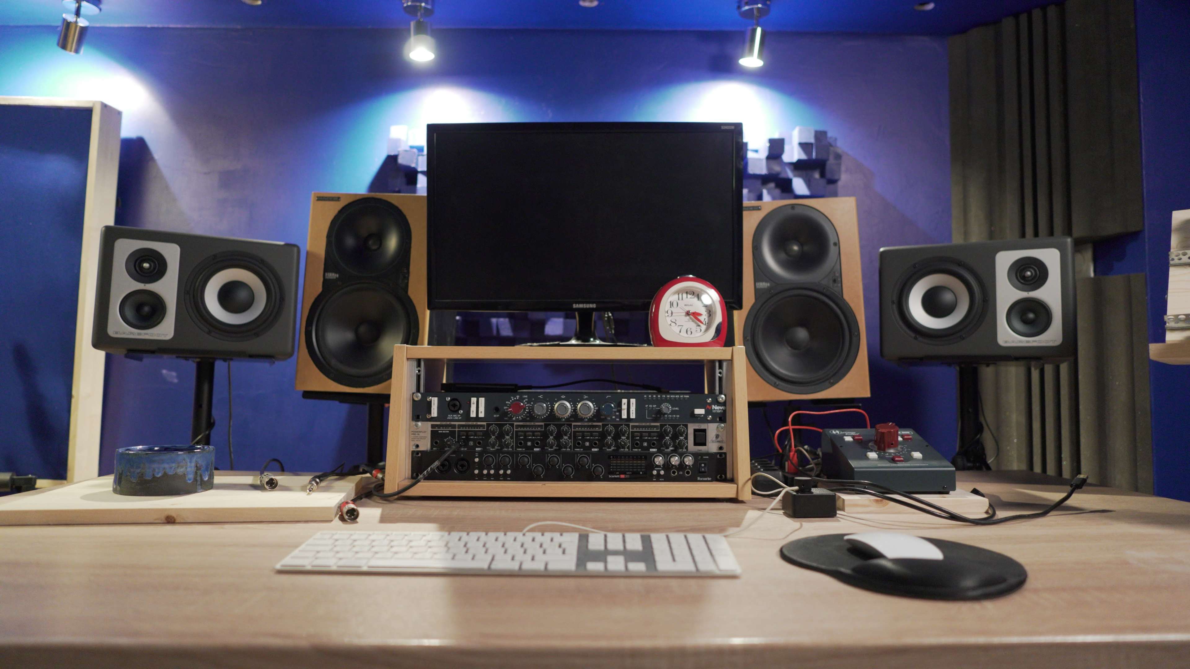 The image shows a music production workspace featuring a computer monitor, audio speakers on both sides, and various audio equipment on a wooden desk.