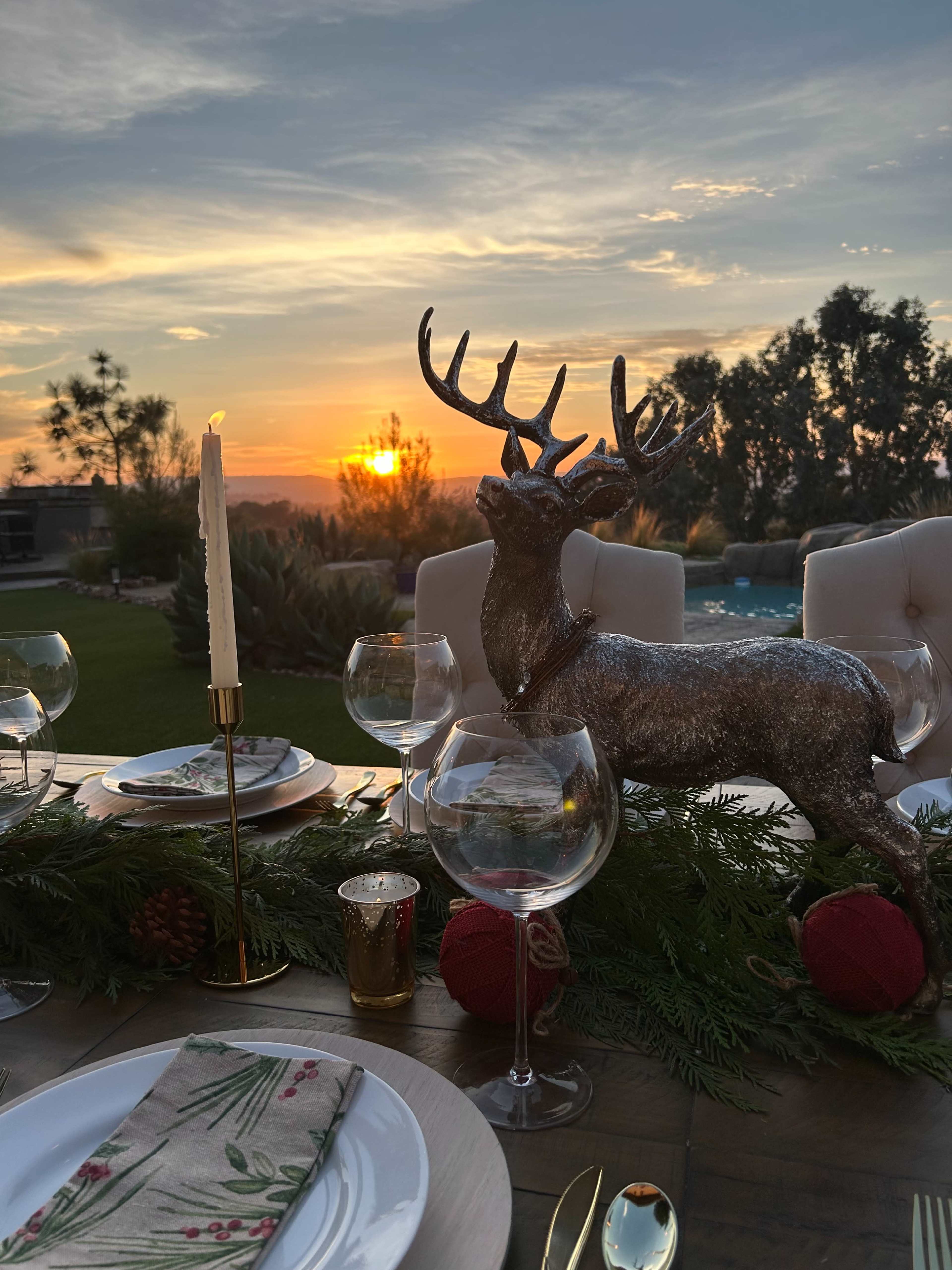 A festive table setting with a decorative reindeer centerpiece, surrounded by wine glasses and green garlands, overlooks a sunset by a pool.