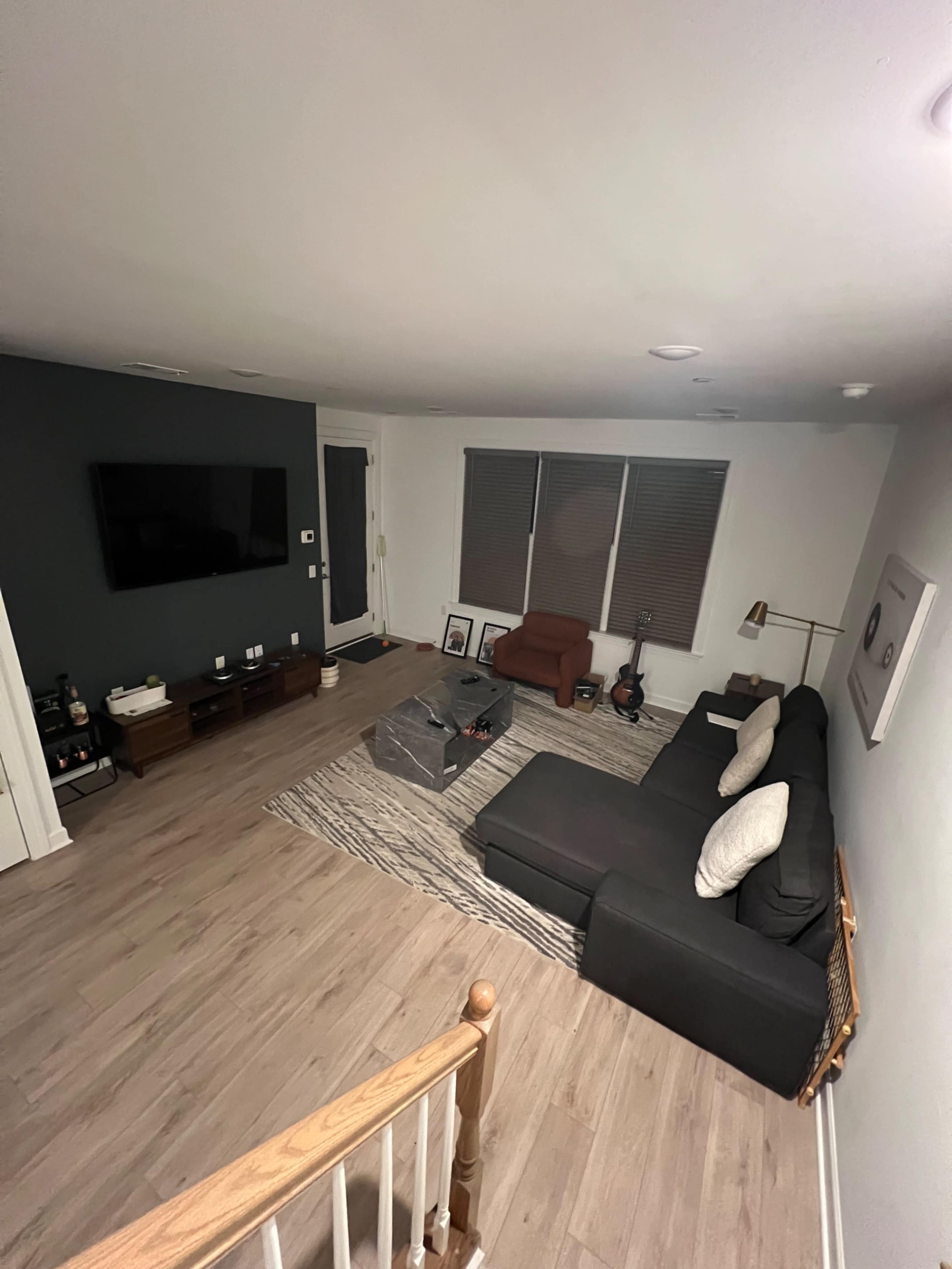 The image shows a modern living room with a sectional sofa, a coffee table, a television mounted on the wall, and natural light coming through large windows.