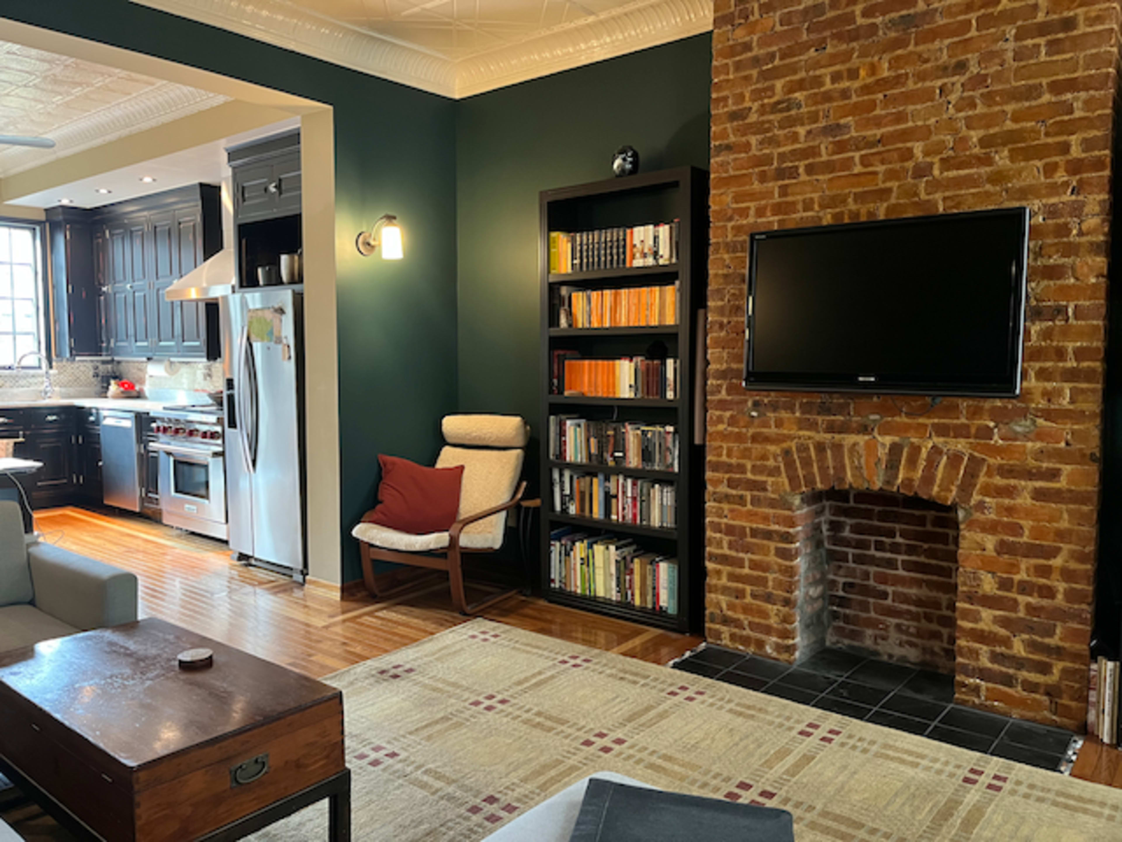 The image shows a cozy living space with a brick accent wall, a bookshelf filled with books, a television mounted above a fireplace, and a kitchen area in the background.