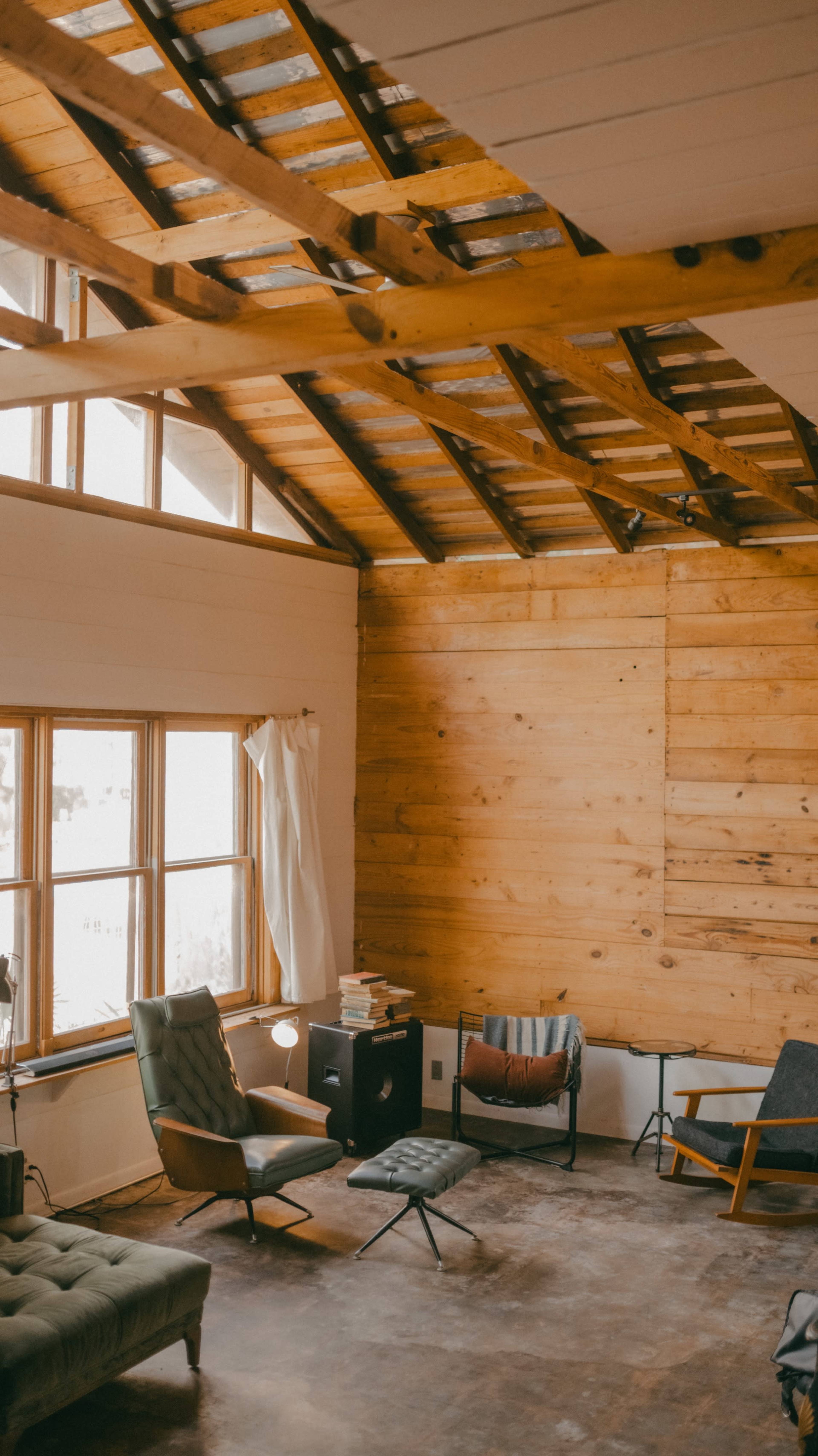 The image shows a spacious interior of a room with wooden walls, large windows, and several chairs arranged in a minimalist style.