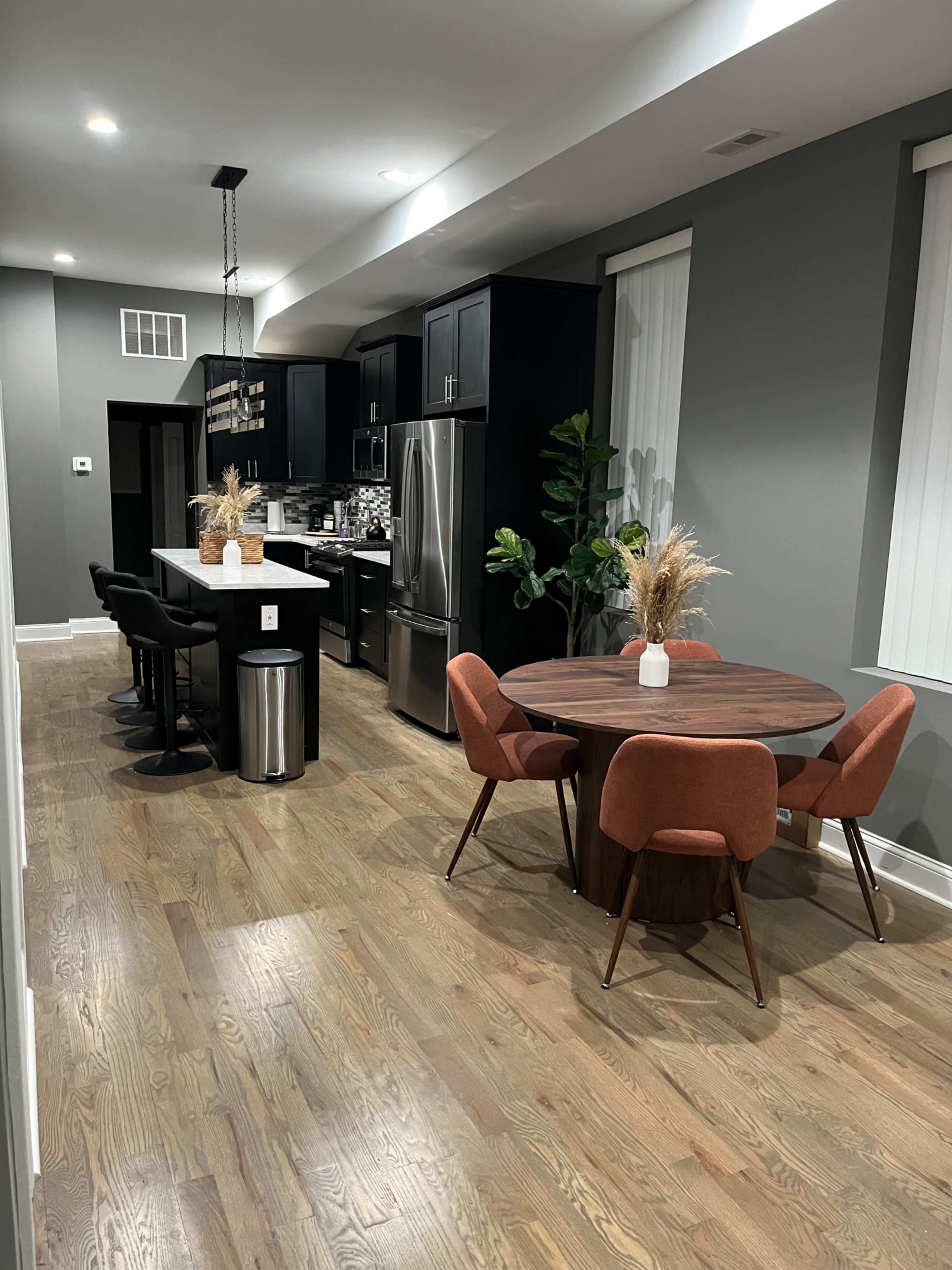 A modern kitchen and dining area featuring black cabinetry, stainless steel appliances, a circular wooden dining table with orange chairs, and a bar with black stools.