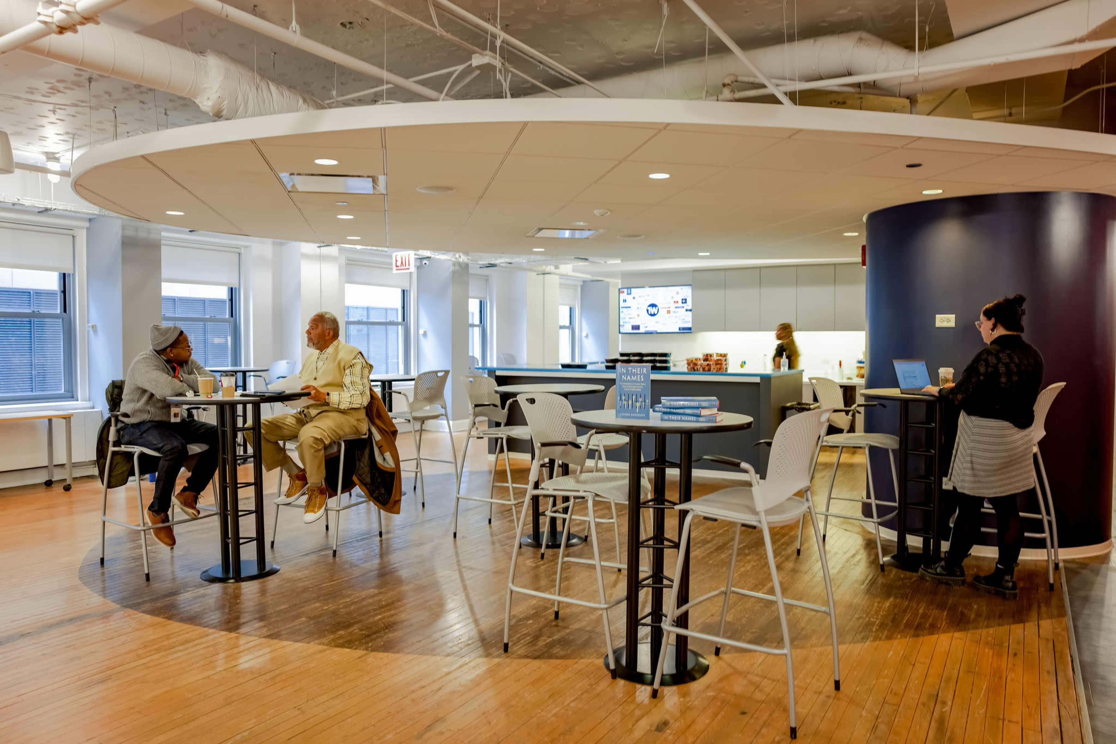 The image shows a modern office space with a circular seating area, featuring high tables and chairs, where a few people are engaged in conversation or working on laptops.
