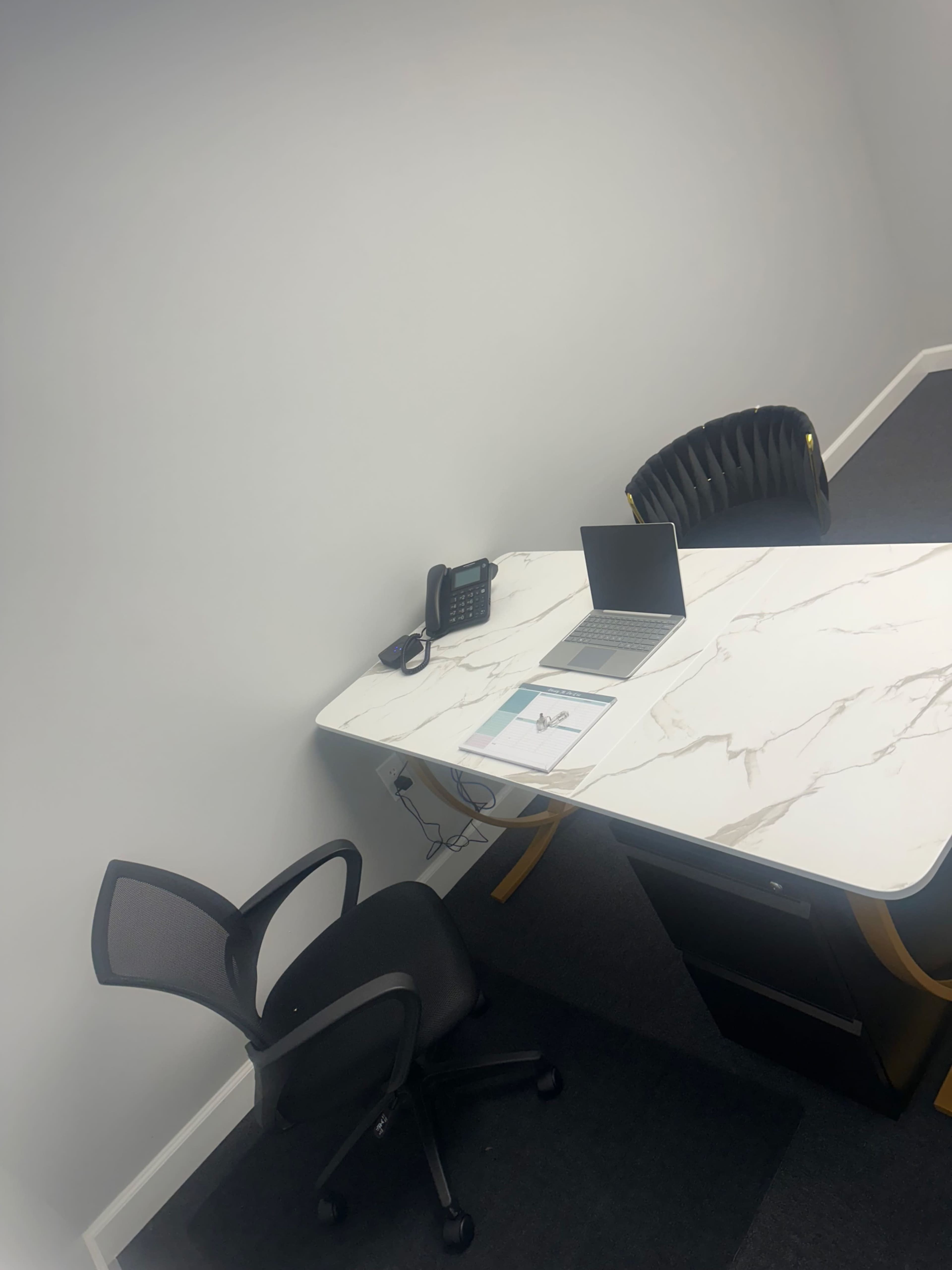A modern office space features a marble-top desk with a laptop, a telephone, and a black office chair, against a plain wall.