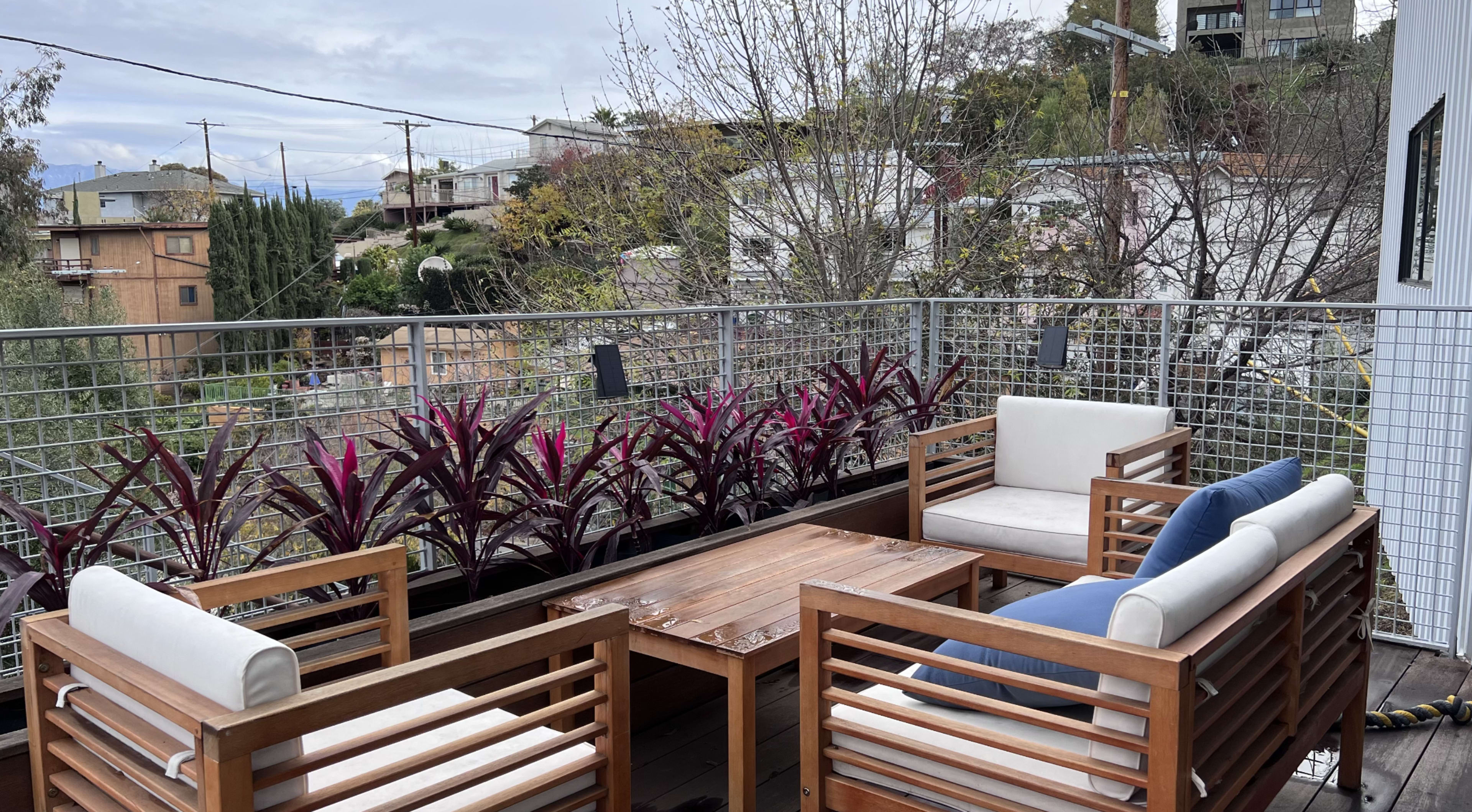 A wooden patio set with white cushions is arranged around a table, surrounded by greenery and purple plants on a balcony.