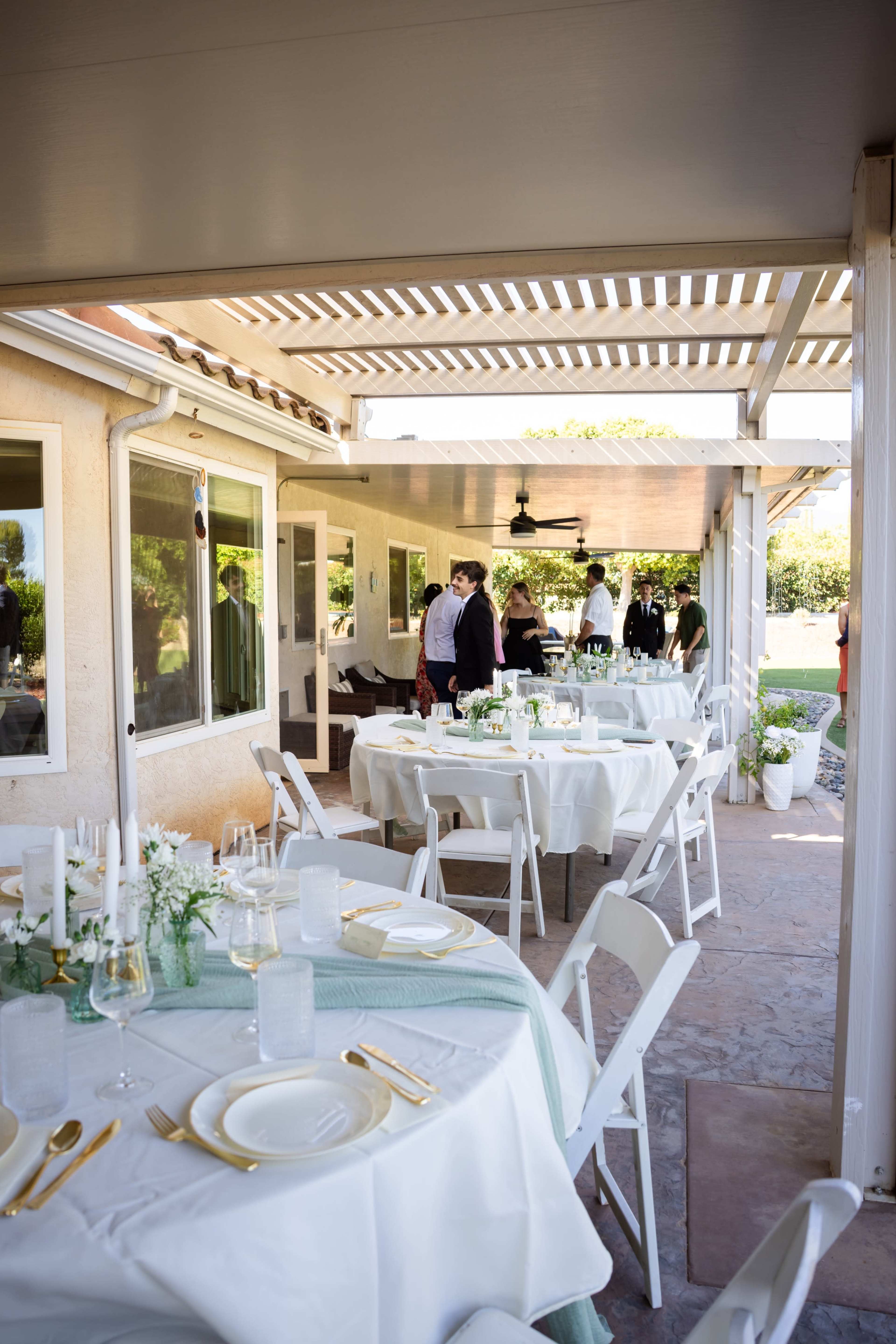 A spacious outdoor patio is arranged with tables set for dining, featuring white tablecloths and elegant place settings, while guests converse in the background.