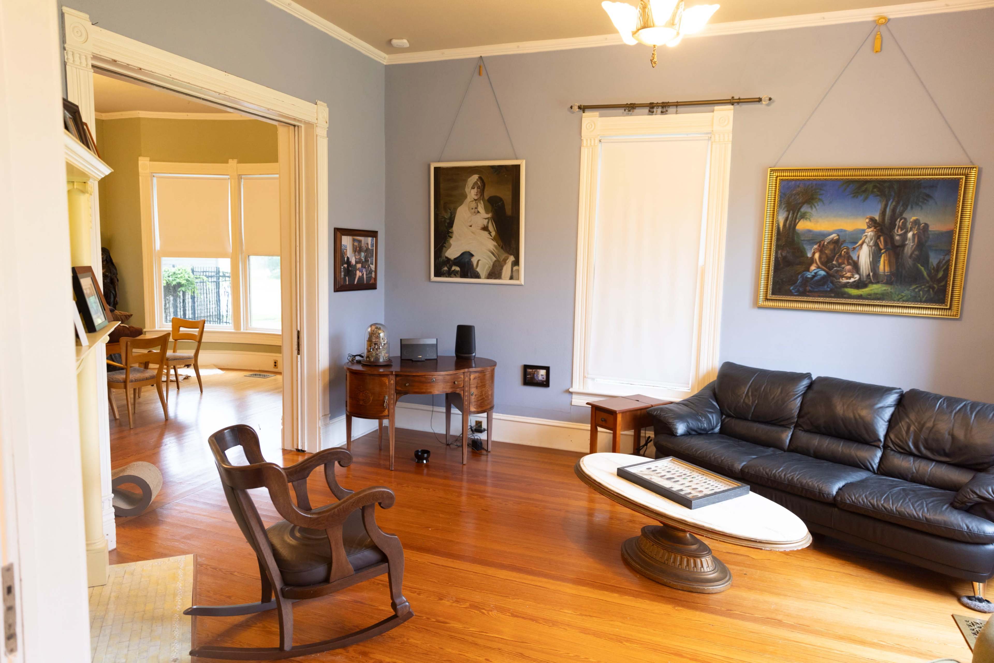 A living room features a black leather sofa, a round coffee table, and various artworks on the walls, with an adjacent dining area visible through an archway.