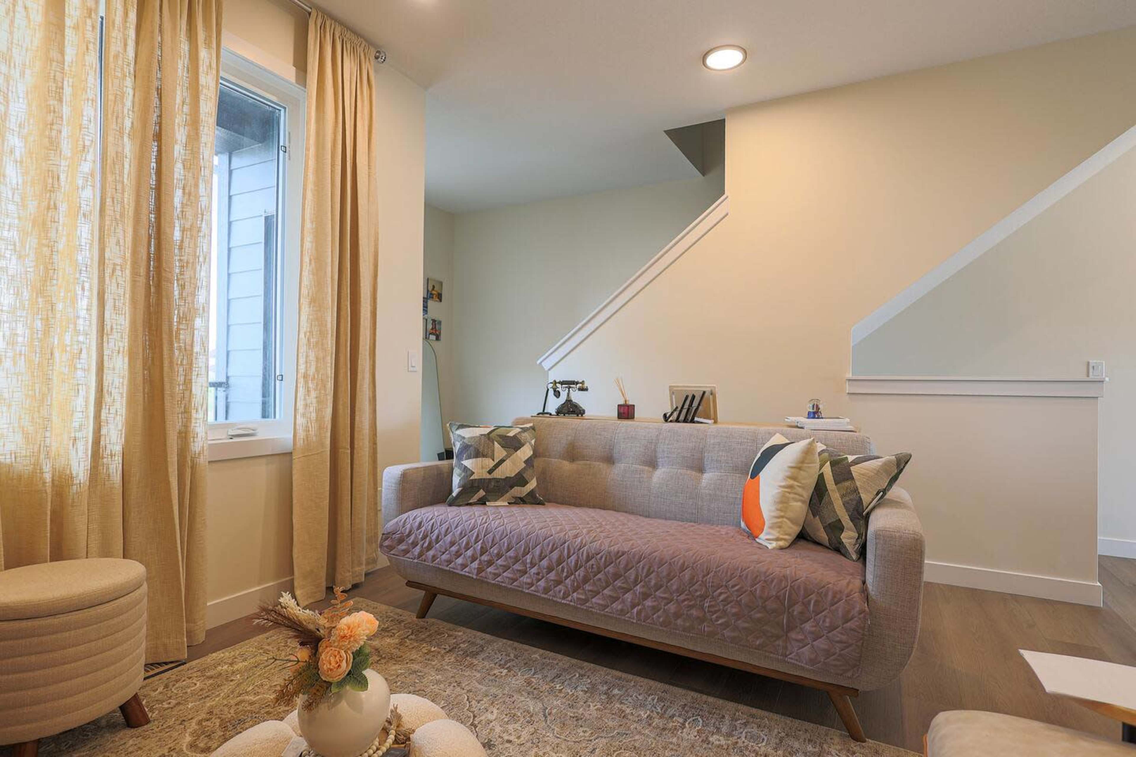 The image shows a cozy living room with a light-colored sofa, decorative pillows, a small coffee table with flowers, and large windows featuring light curtains.