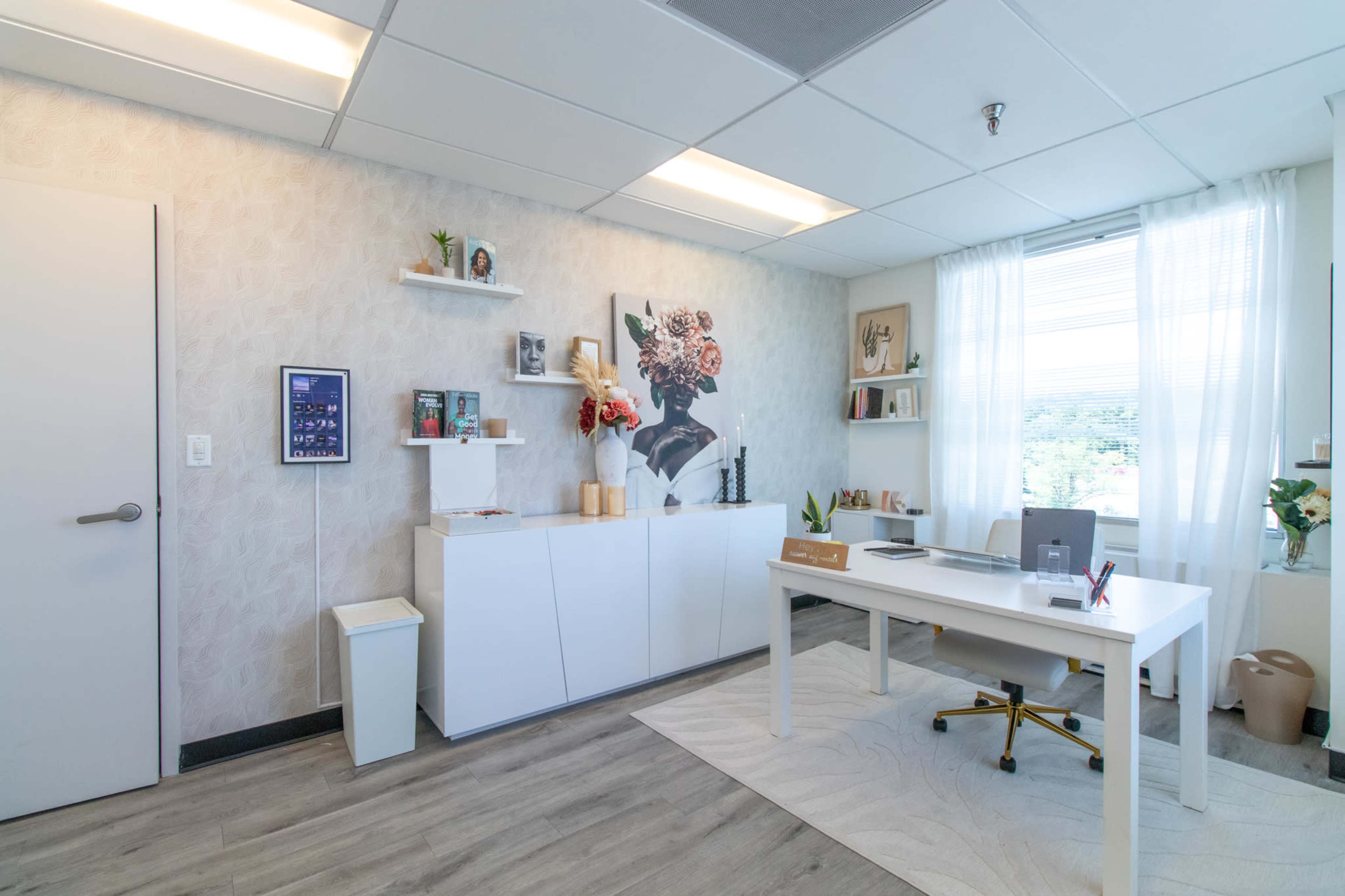 A well-organized office area features a white desk, a minimalistic shelf with decorative items, and a large window with sheer curtains letting in natural light.