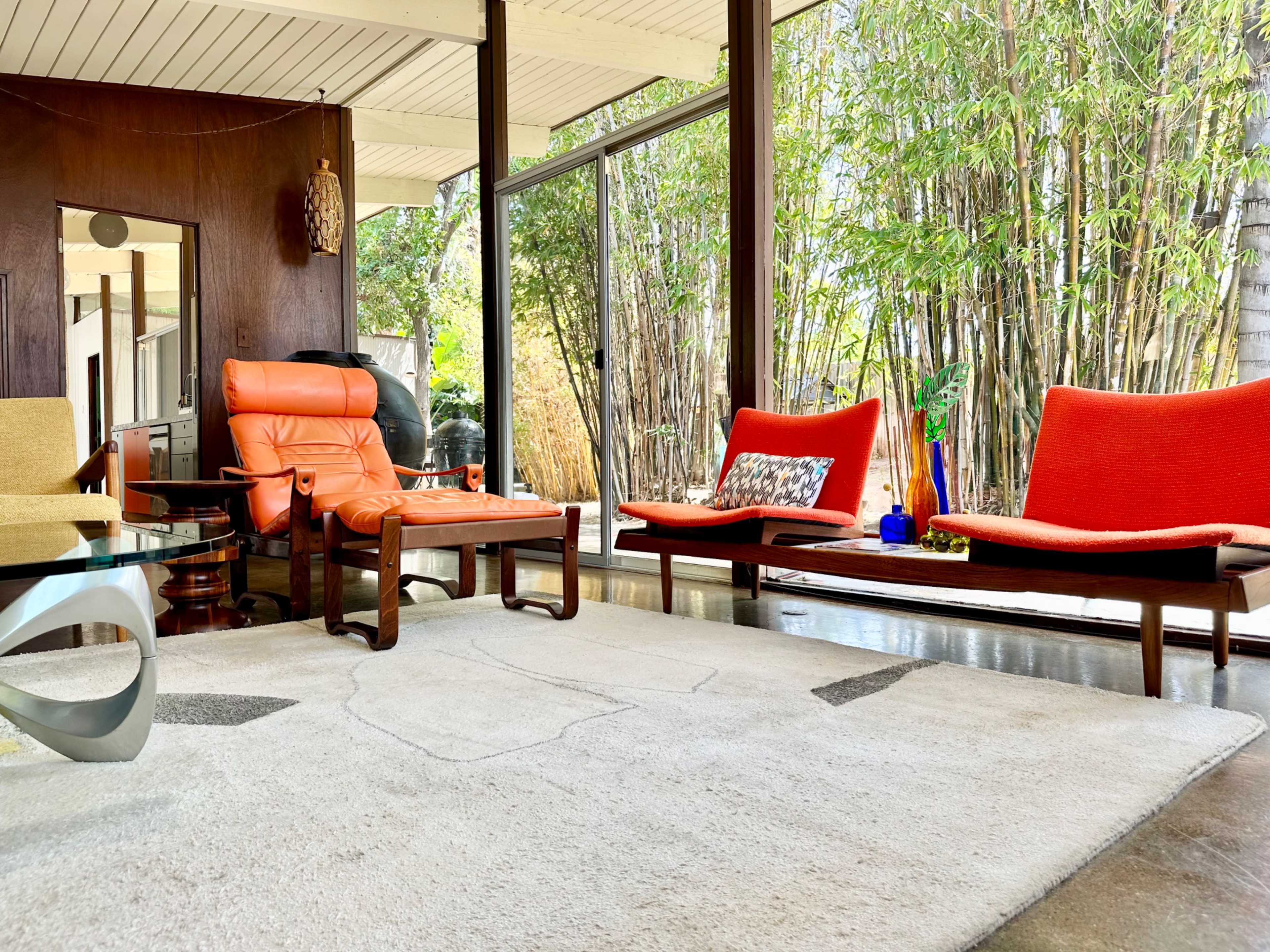 The scene features a modern living room with orange and red furniture, large windows overlooking bamboo plants, and a light-colored rug on a polished floor.