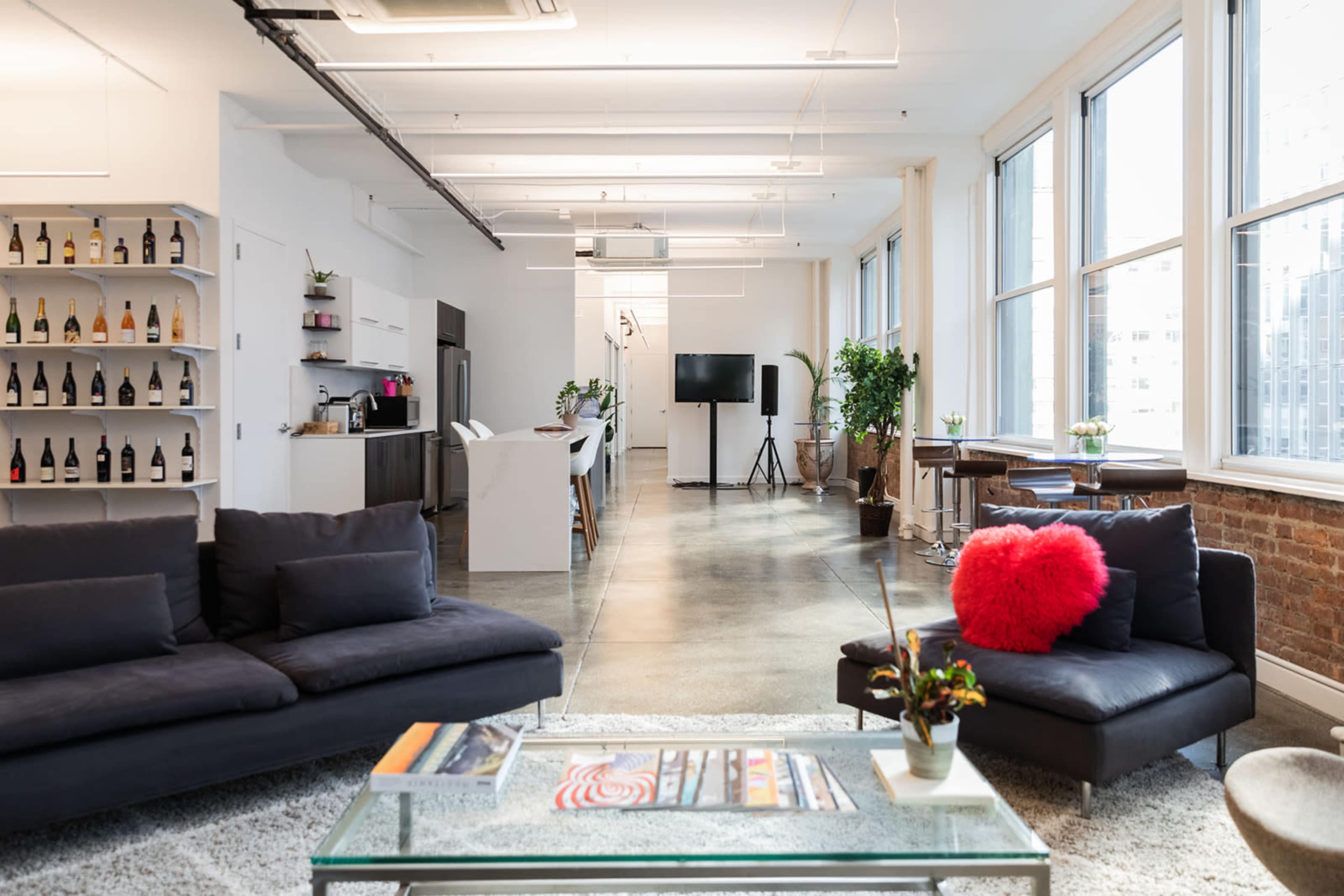 A modern open-concept office space featuring a lounge area with dark sofas, a glass coffee table, and a kitchen area with shelves holding various bottles.