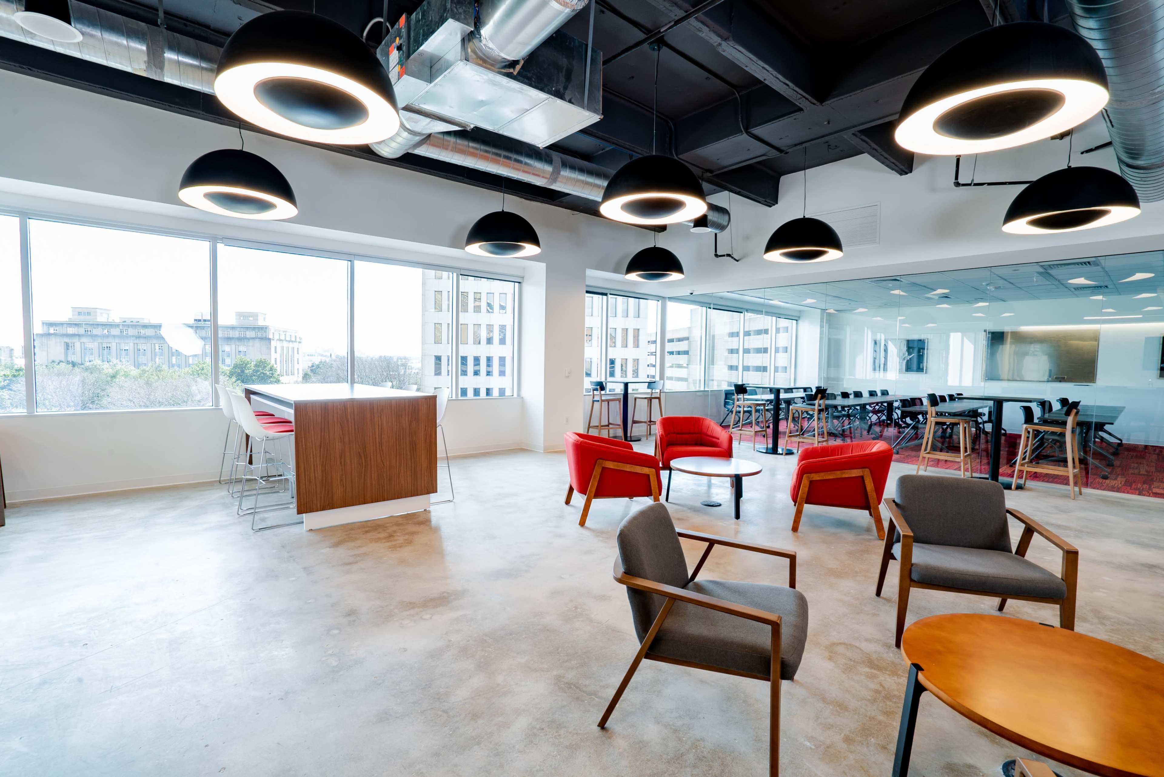 The image shows a modern office space featuring a set of chairs and tables, large windows allowing natural light, and hanging circular lights.