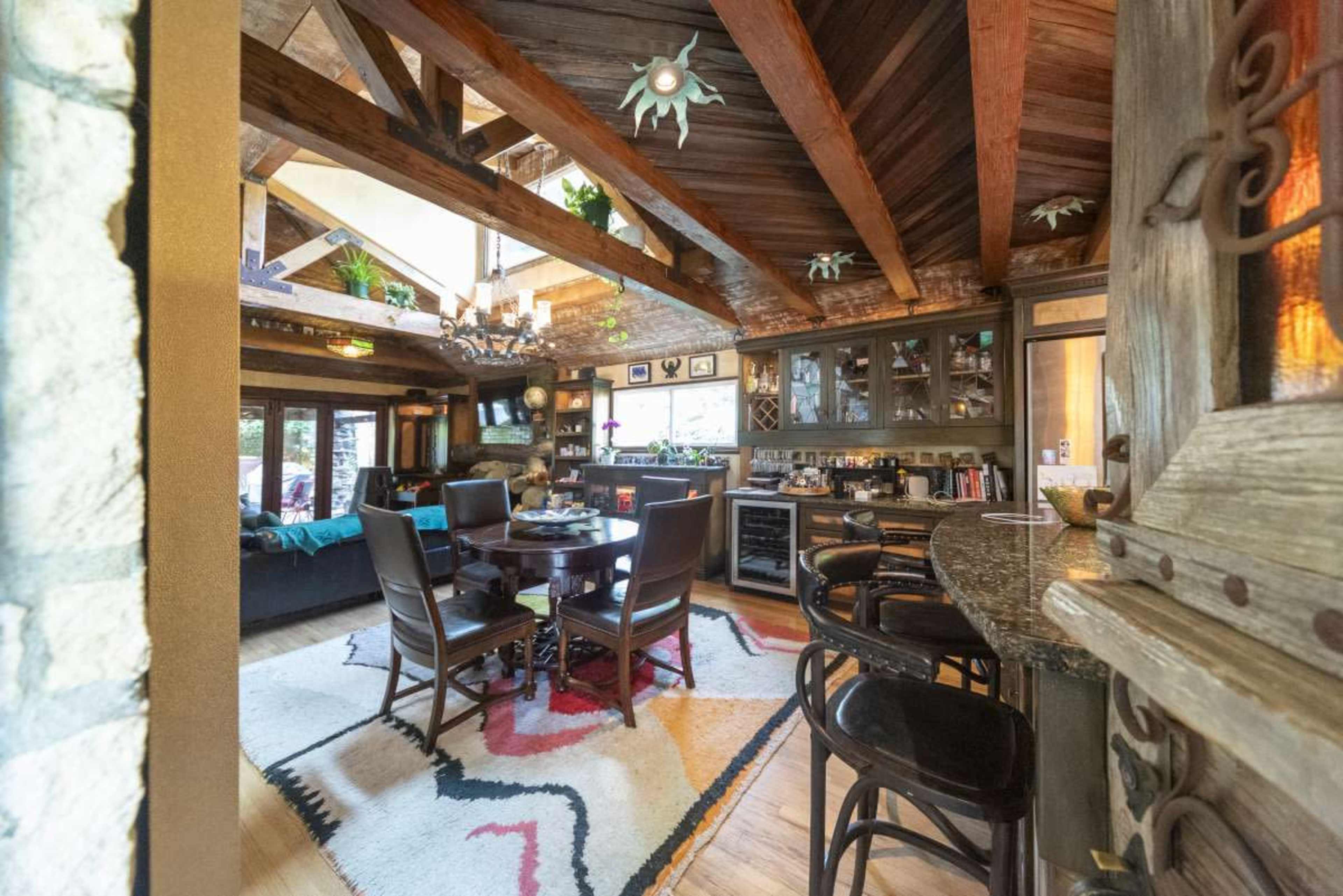 The image shows a rustic kitchen and dining area featuring wooden beams, a circular table with chairs, and a cozy seating area with a sofa.