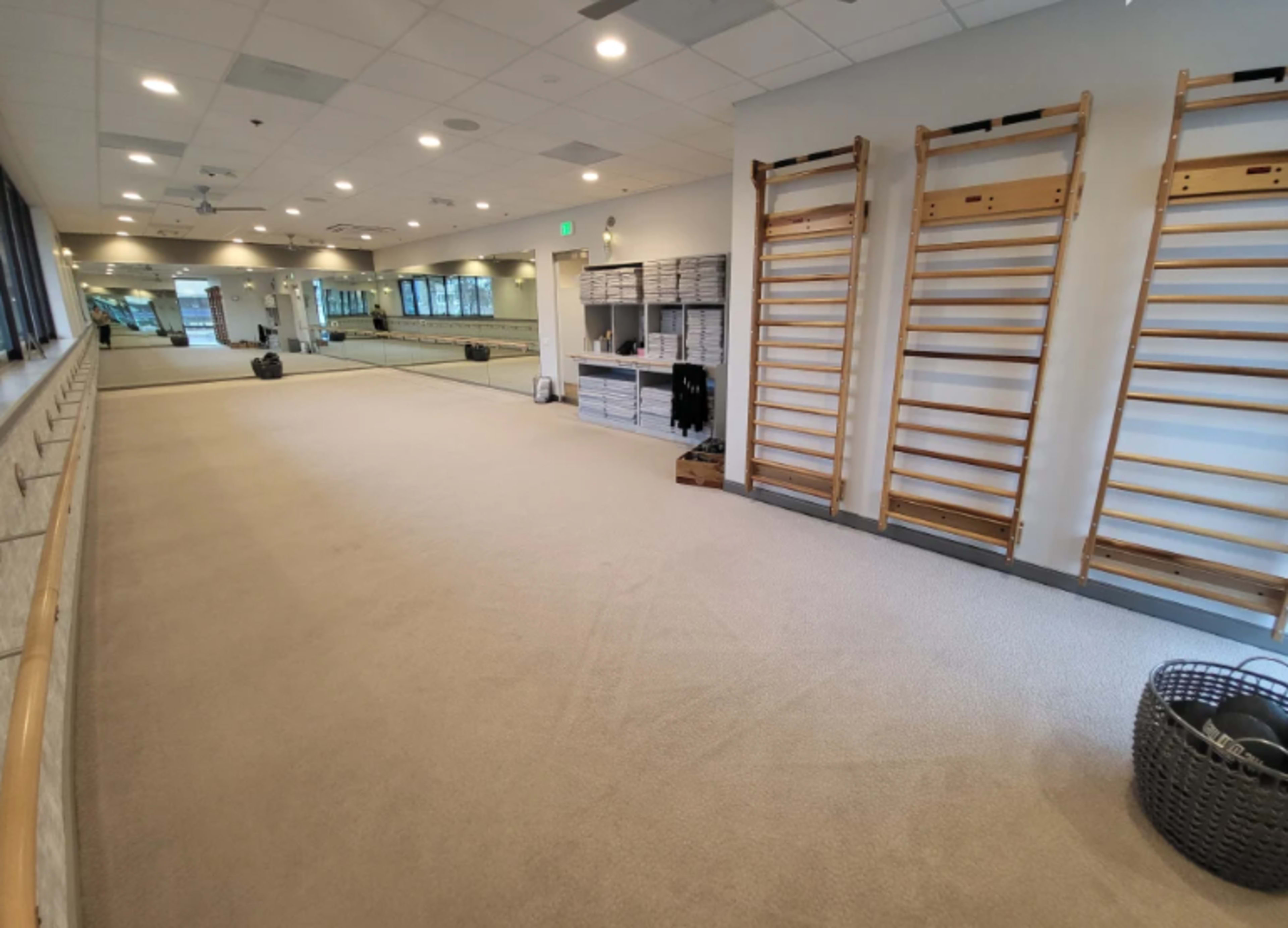 The image shows a spacious exercise studio with wooden wall bars, a large mirror, and neatly organized storage cubicles.
