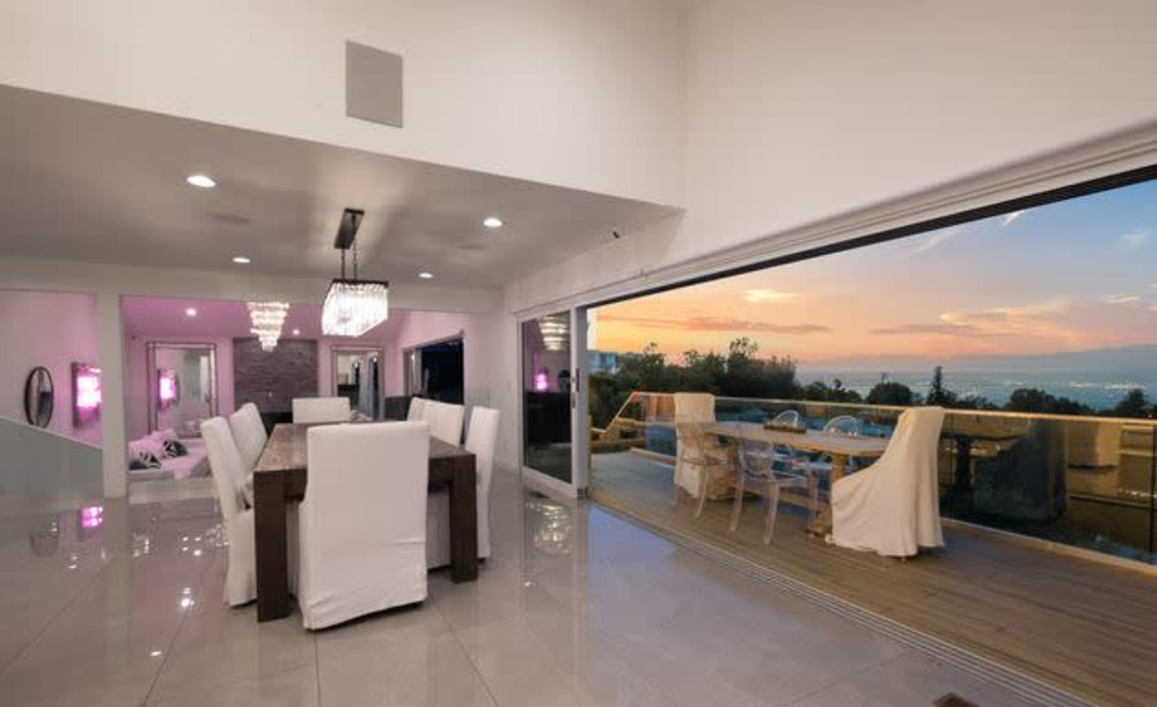 The image shows a modern dining area with a large table and white chairs, featuring a glass door that opens to a patio with outdoor seating and a view of the sunset.