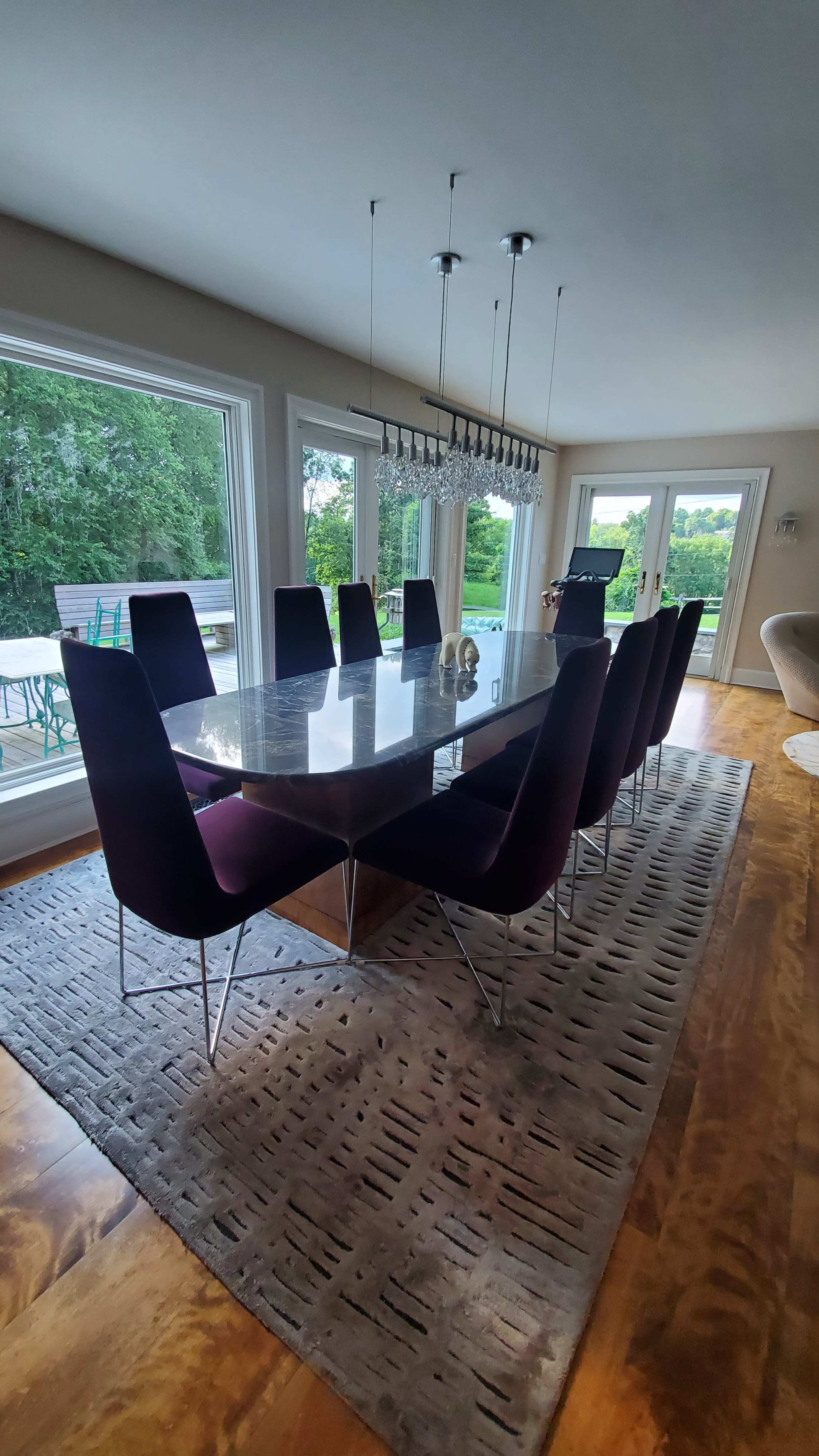 A long dining table with purple chairs is positioned in a spacious room featuring large windows that overlook a green landscape.