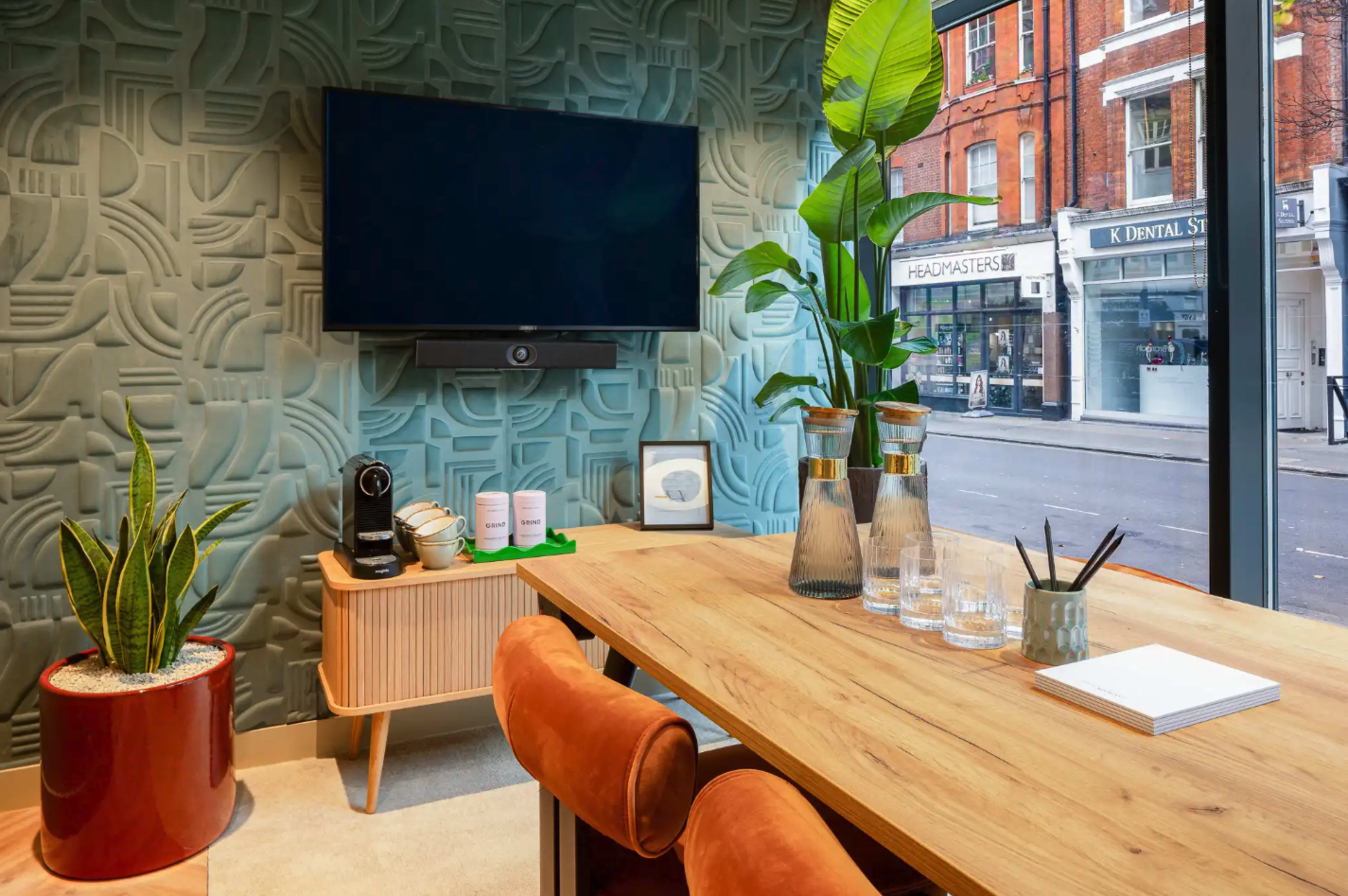 The image shows a modern office space featuring a wooden table with orange chairs, a large television mounted on a textured green wall, and decorative plants beside a window overlooking a street.