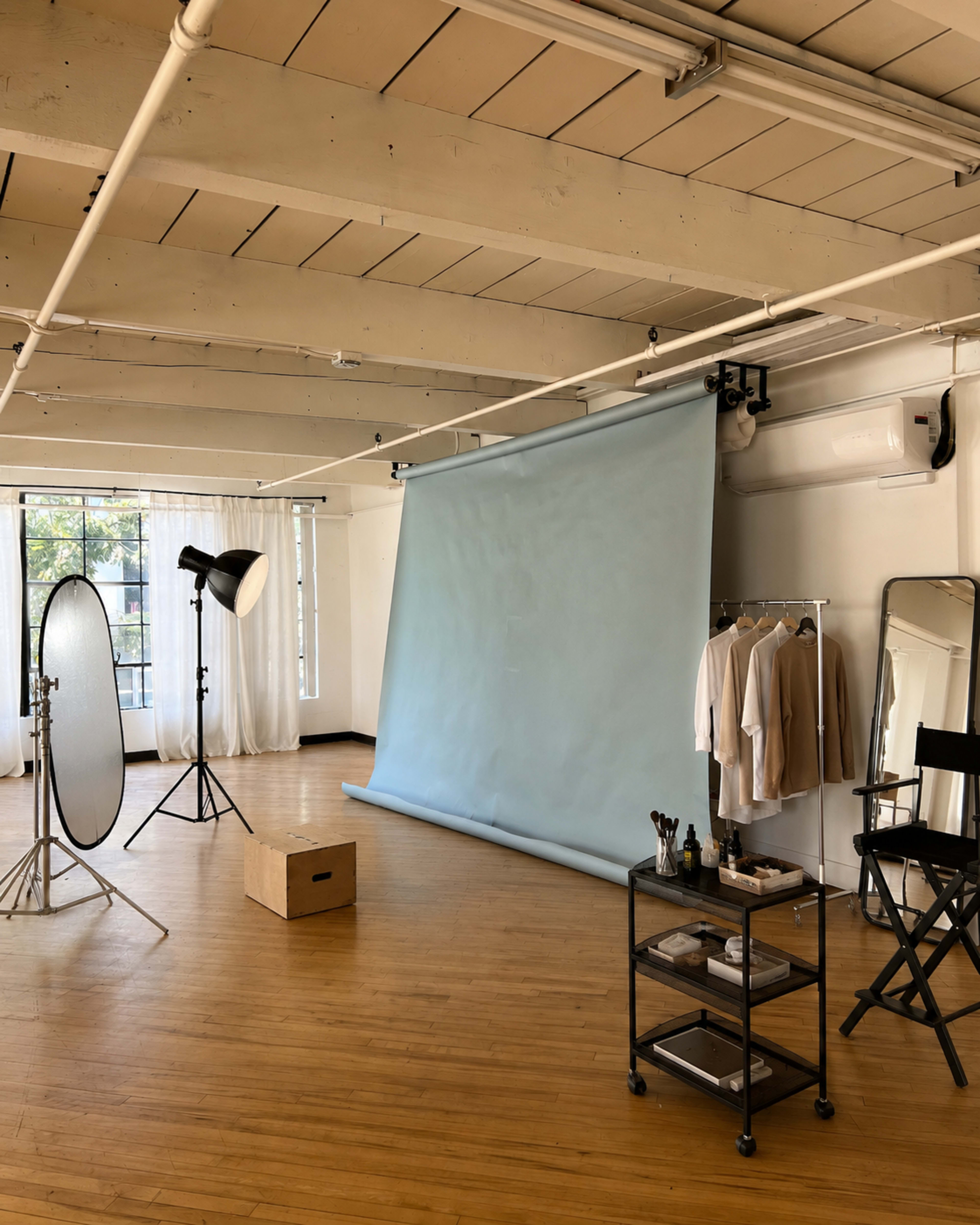 The image shows a well-lit studio space with a light blue backdrop, a softbox light, a clothing rack with beige attire, a mirror, a director's chair, and a cart with makeup tools.