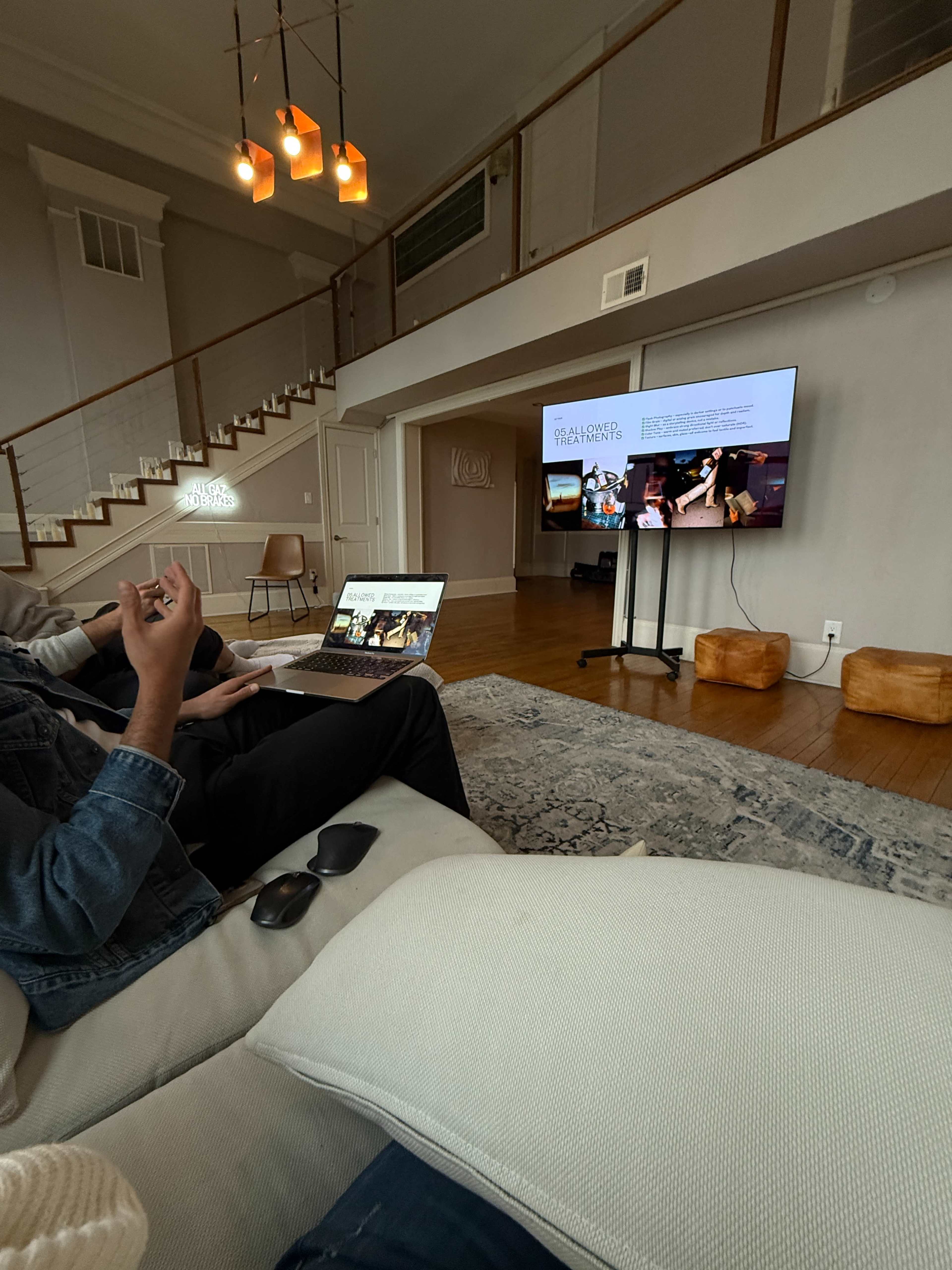 A person is seated on a couch, using a laptop, while facing a large television displaying images and text in a well-lit living room.