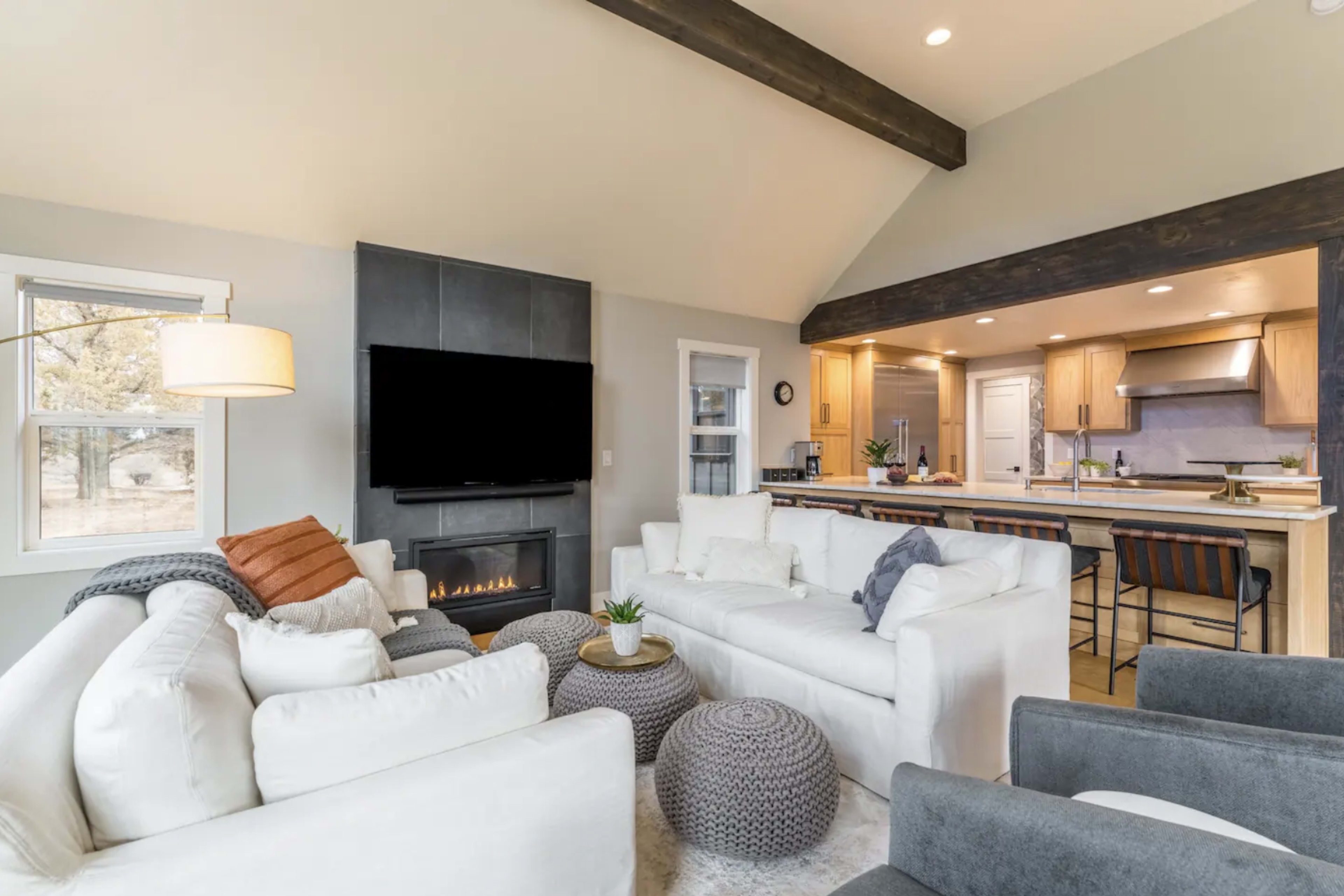 A modern living room features white sofas arranged around a coffee table, with a television mounted on the gray wall and a kitchen visible in the background.