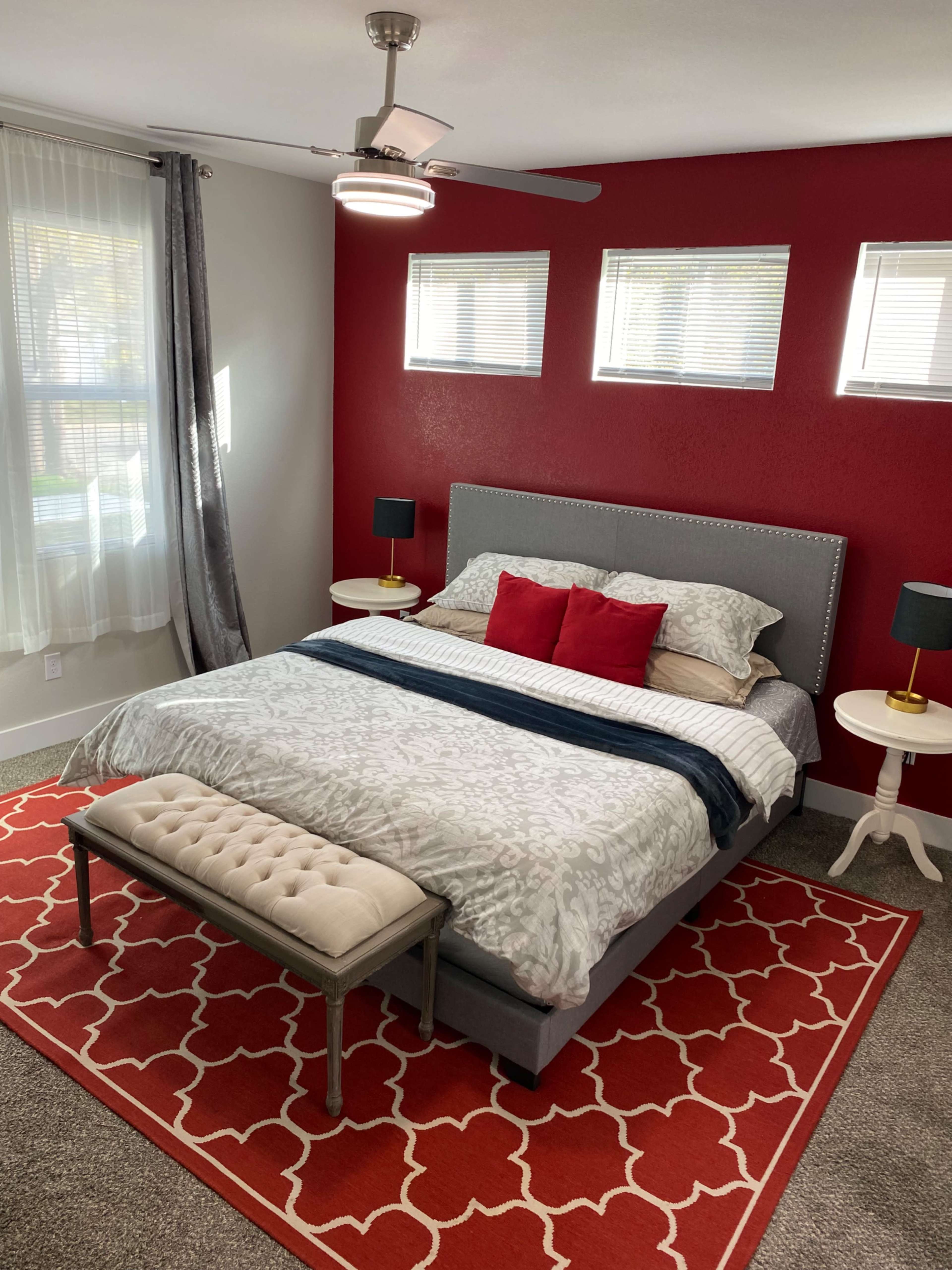 The image shows a well-decorated bedroom featuring a gray bed with white and red bedding, red accent walls, and stylish furniture including a bench and side tables.