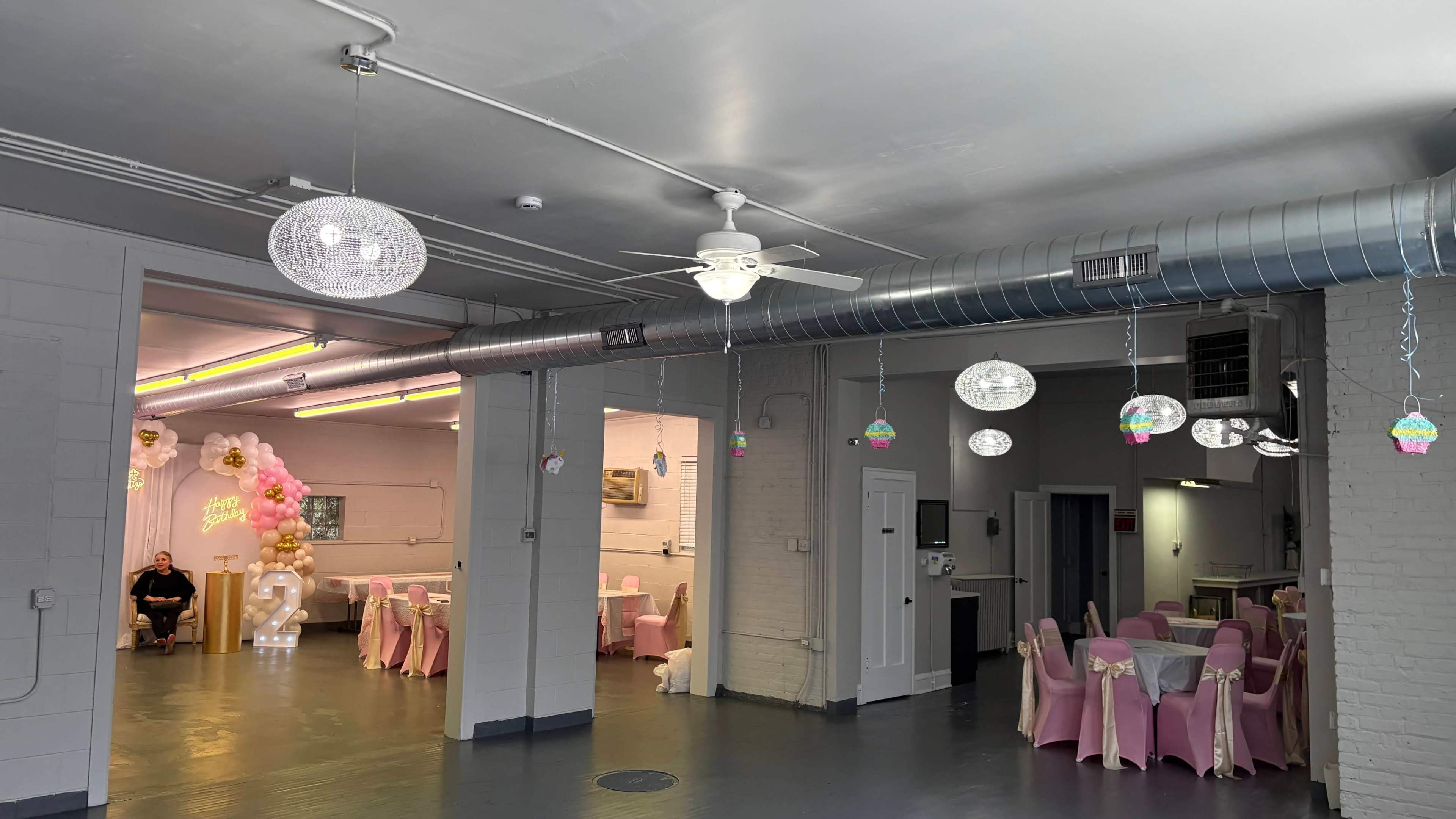 The image shows a spacious event hall decorated with pastel-colored balloons and tables set with pink cloths, featuring hanging light fixtures and an organized seating area.