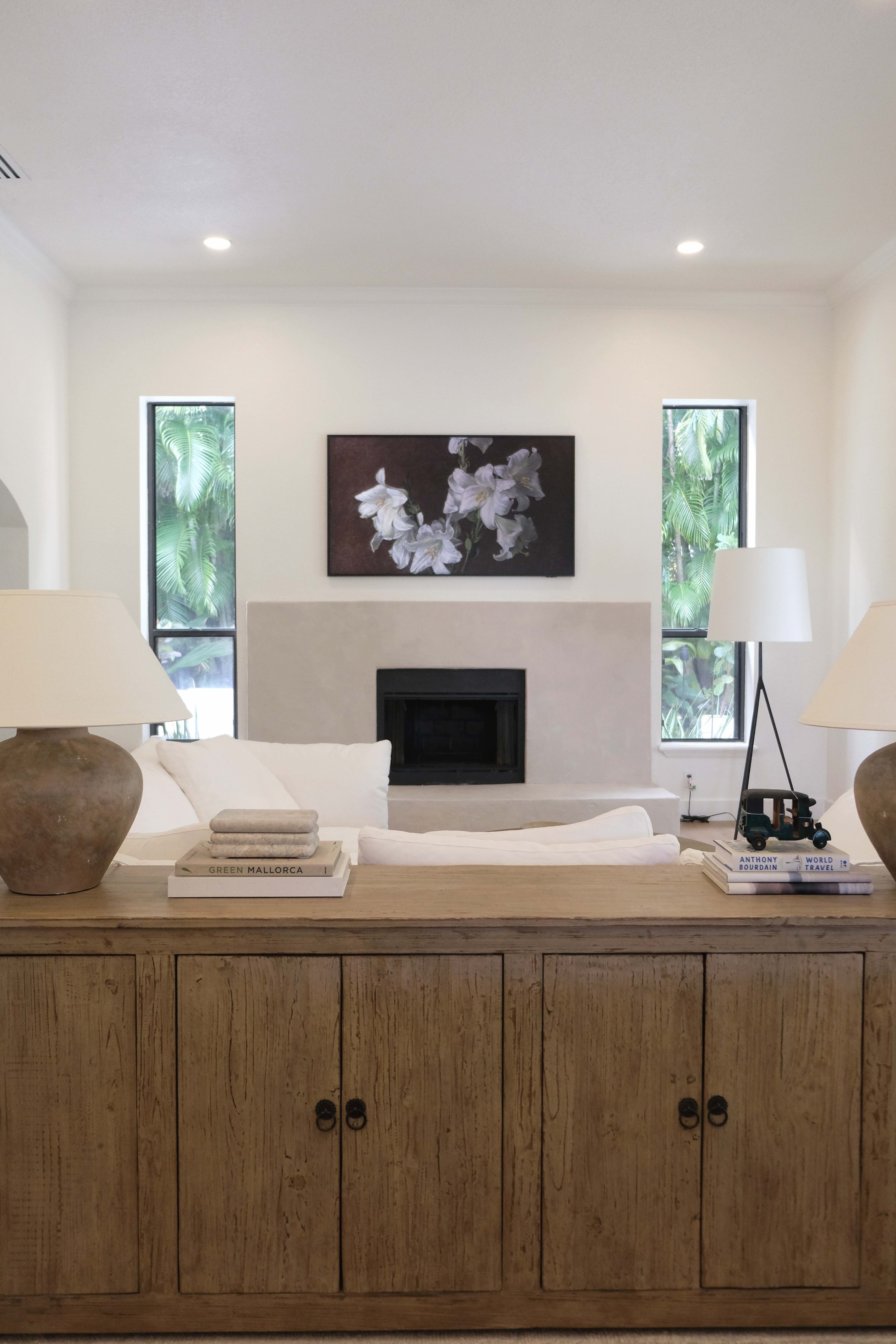 The image shows a modern living room with a white sofa, lamps on either side of a wooden console table, and a fireplace beneath a piece of wall art featuring flowers.