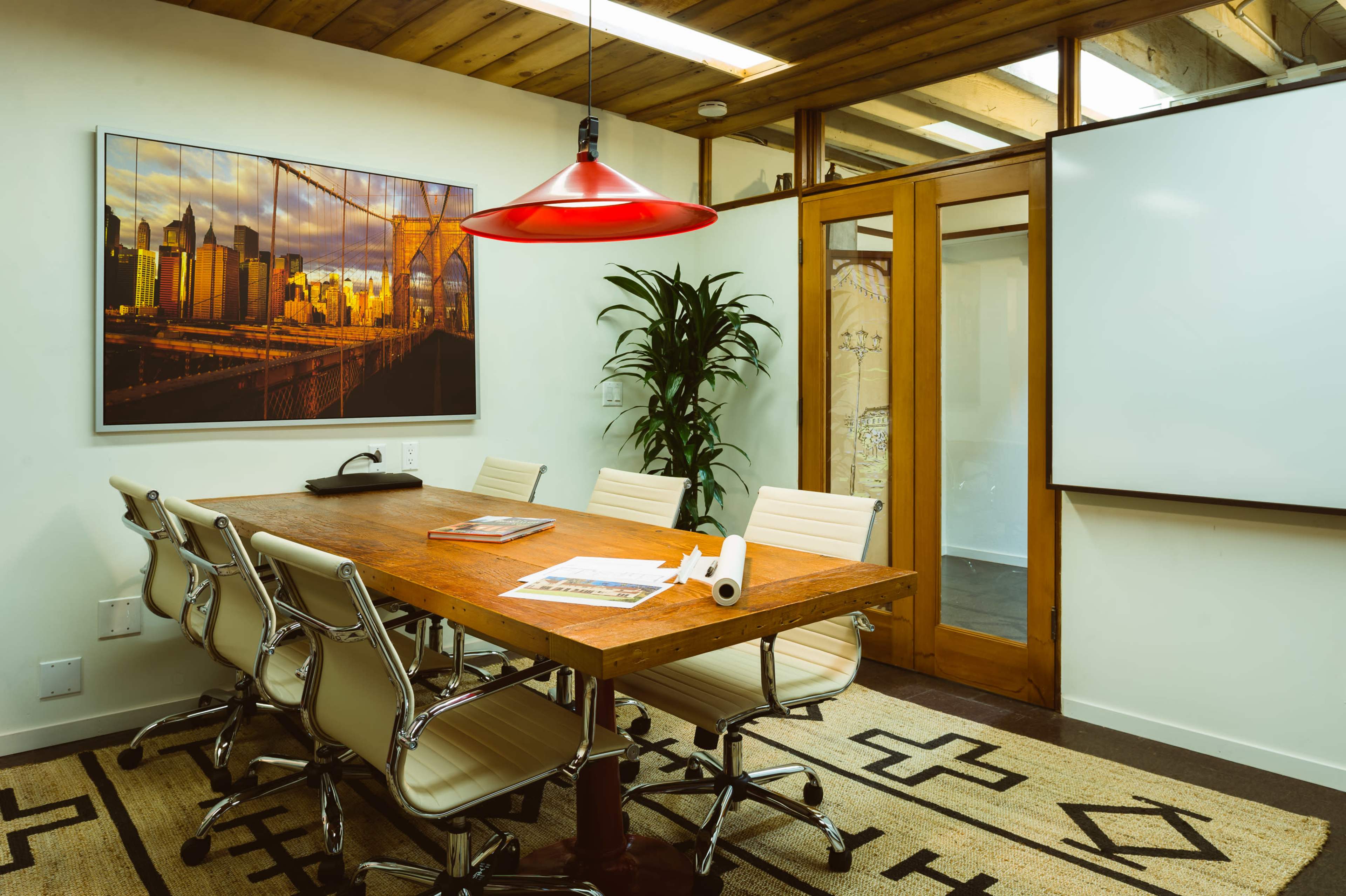 A modern conference room features a wooden table surrounded by white chairs, with a large artwork on the wall and a red pendant light overhead.
