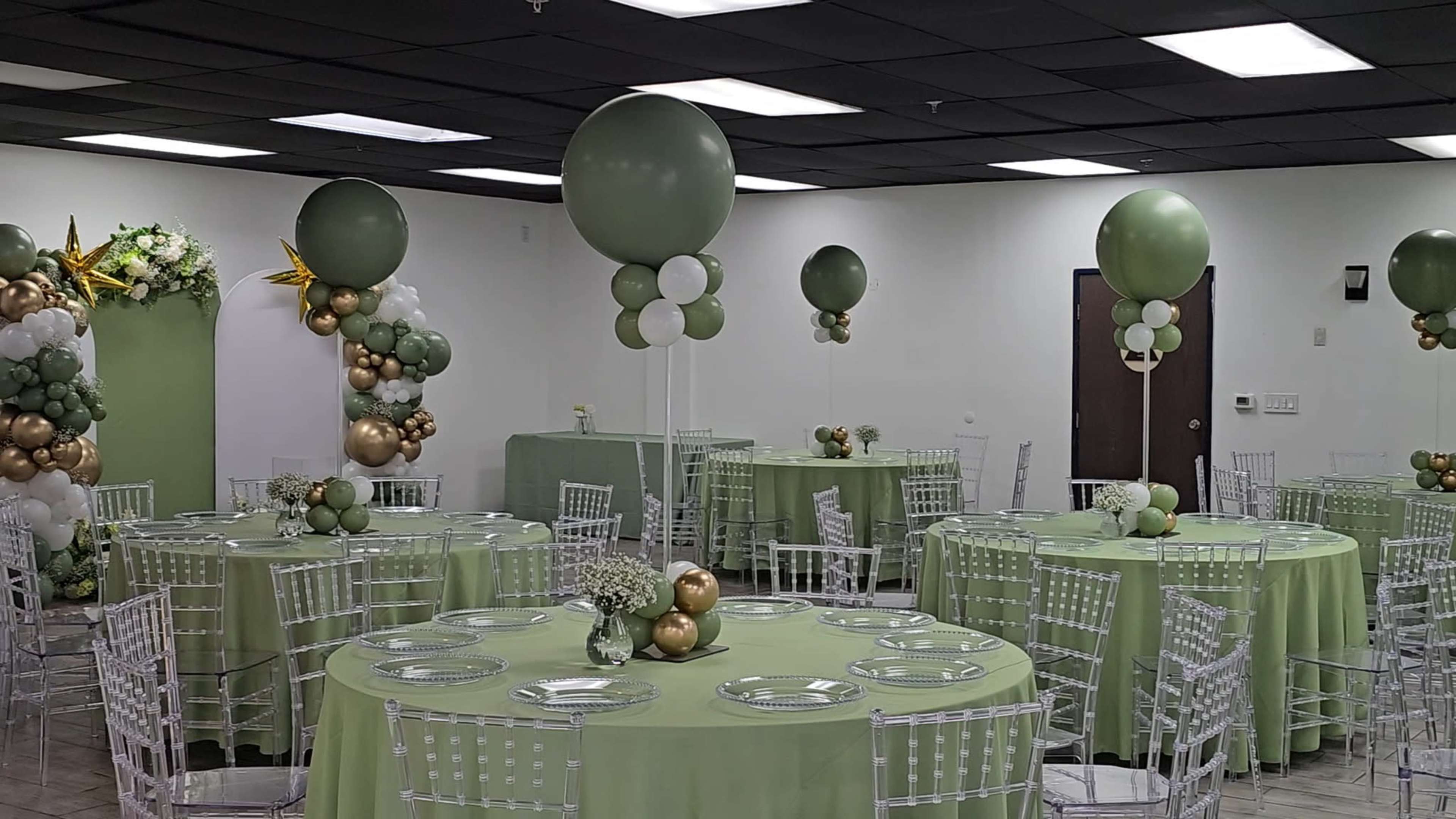 The image shows a banquet hall decorated with green and gold balloons, circular tables covered in green tablecloths, and transparent Chiavari chairs.