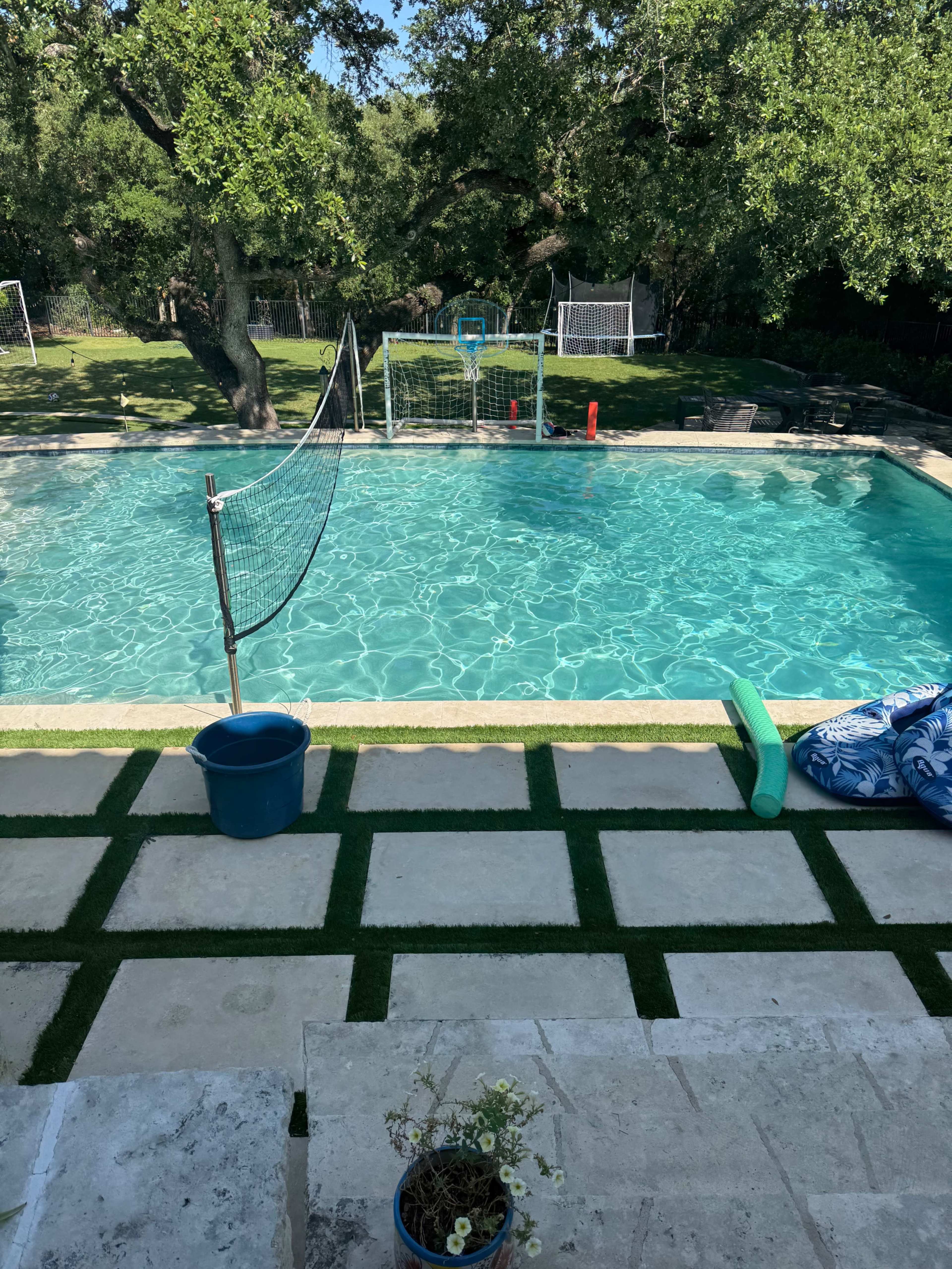 A backyard swimming pool is surrounded by green grass and a volleyball net, with lounge chairs and a potted plant nearby.
