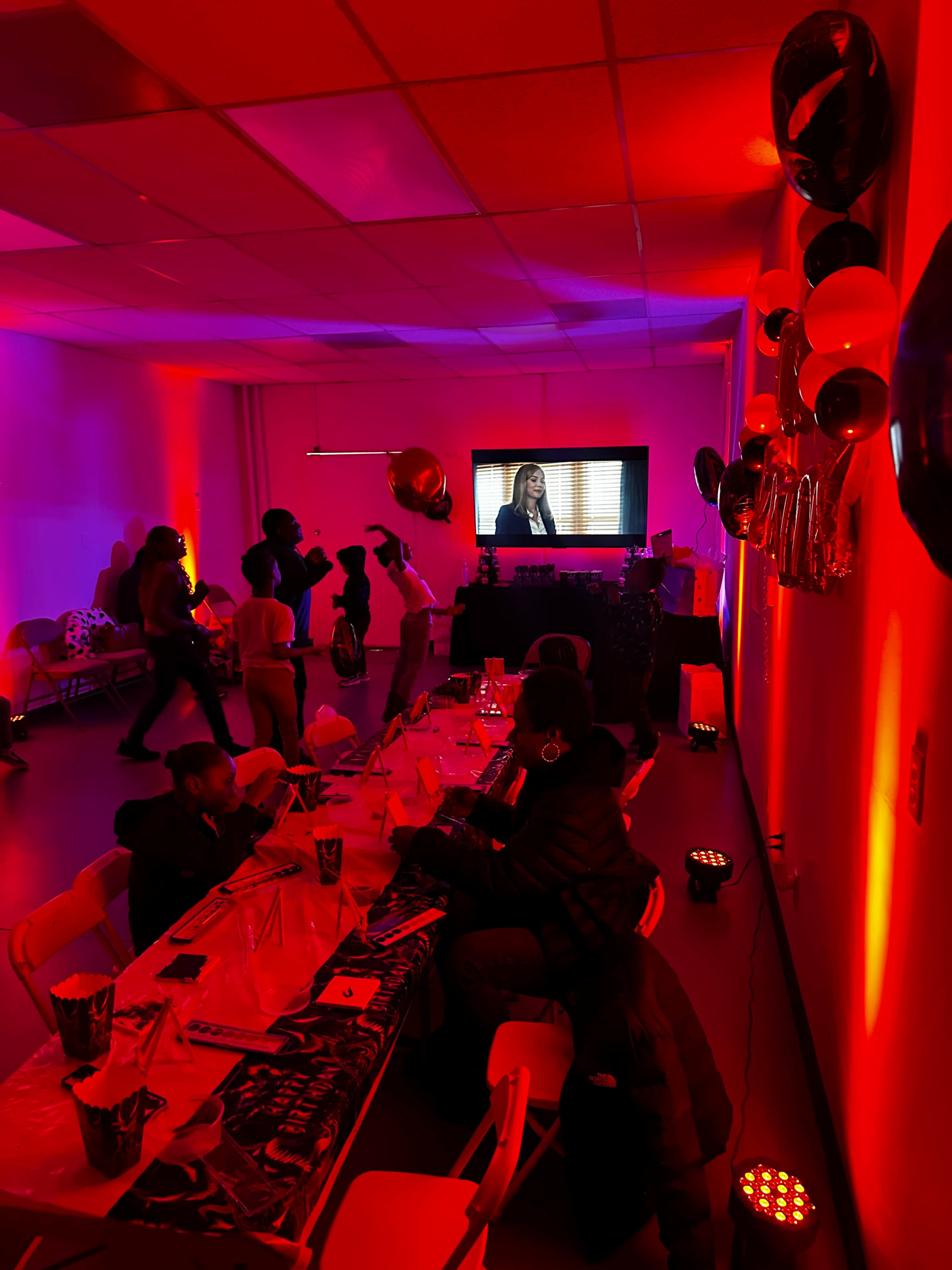 A gathering takes place in a dimly lit room with red lighting, featuring a table set for dining and a screen displaying a video.