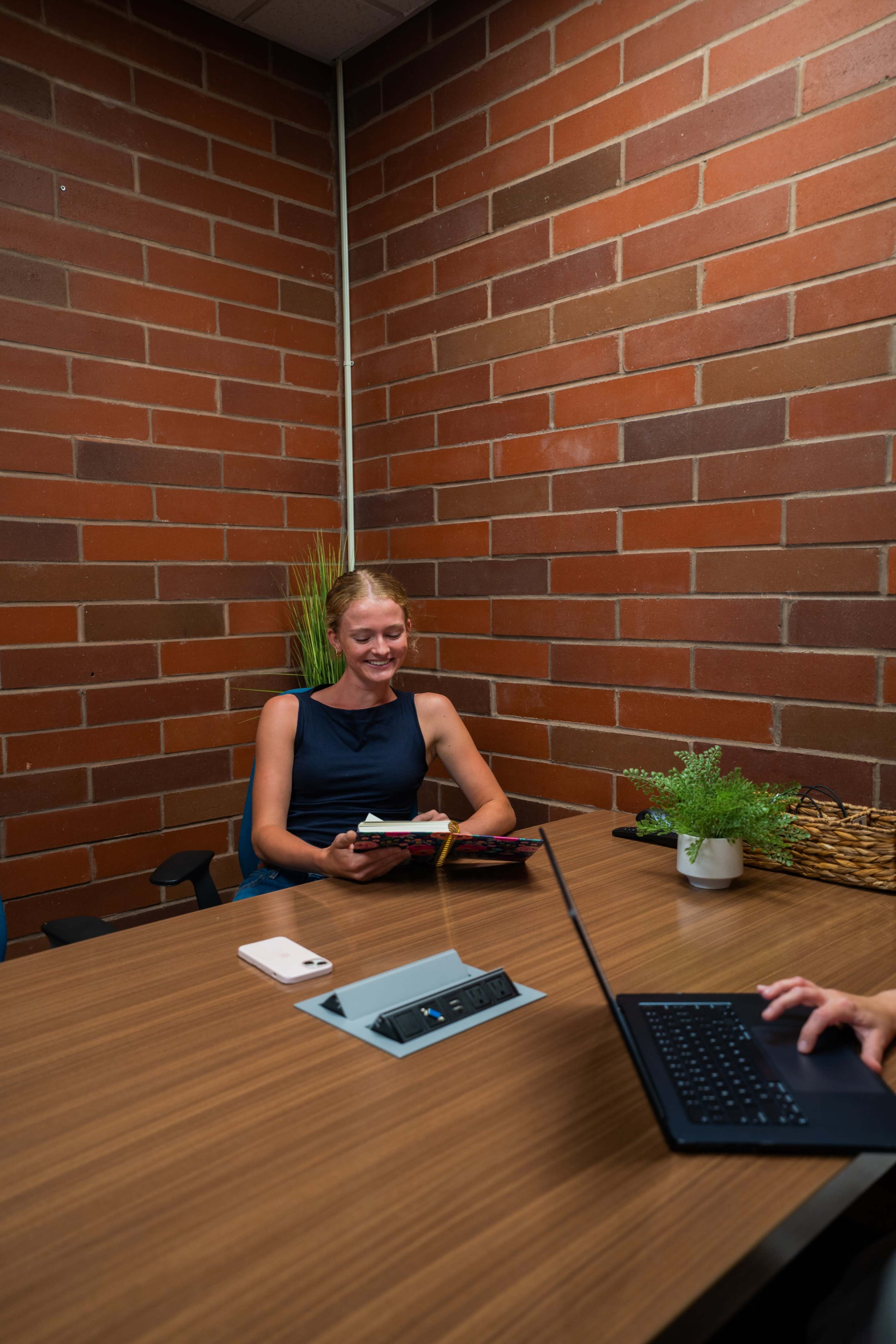 A woman sits at a table in a small meeting room with brick walls, holding a notebook while smiling at another person using a laptop.
