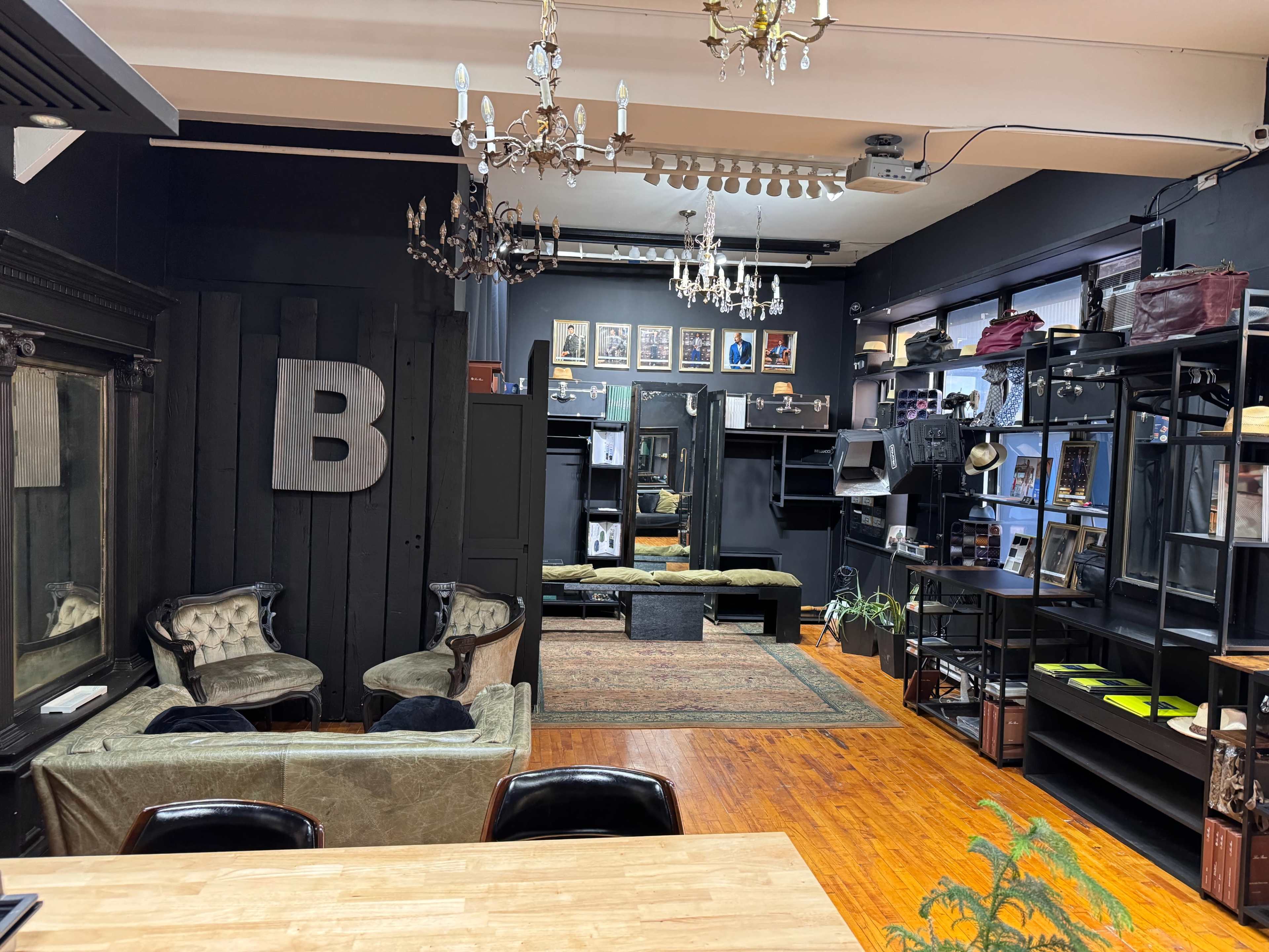 The interior of a stylish salon features dark walls, chandeliers, and a mix of vintage and modern furniture, with a large letter "B" on one wall.