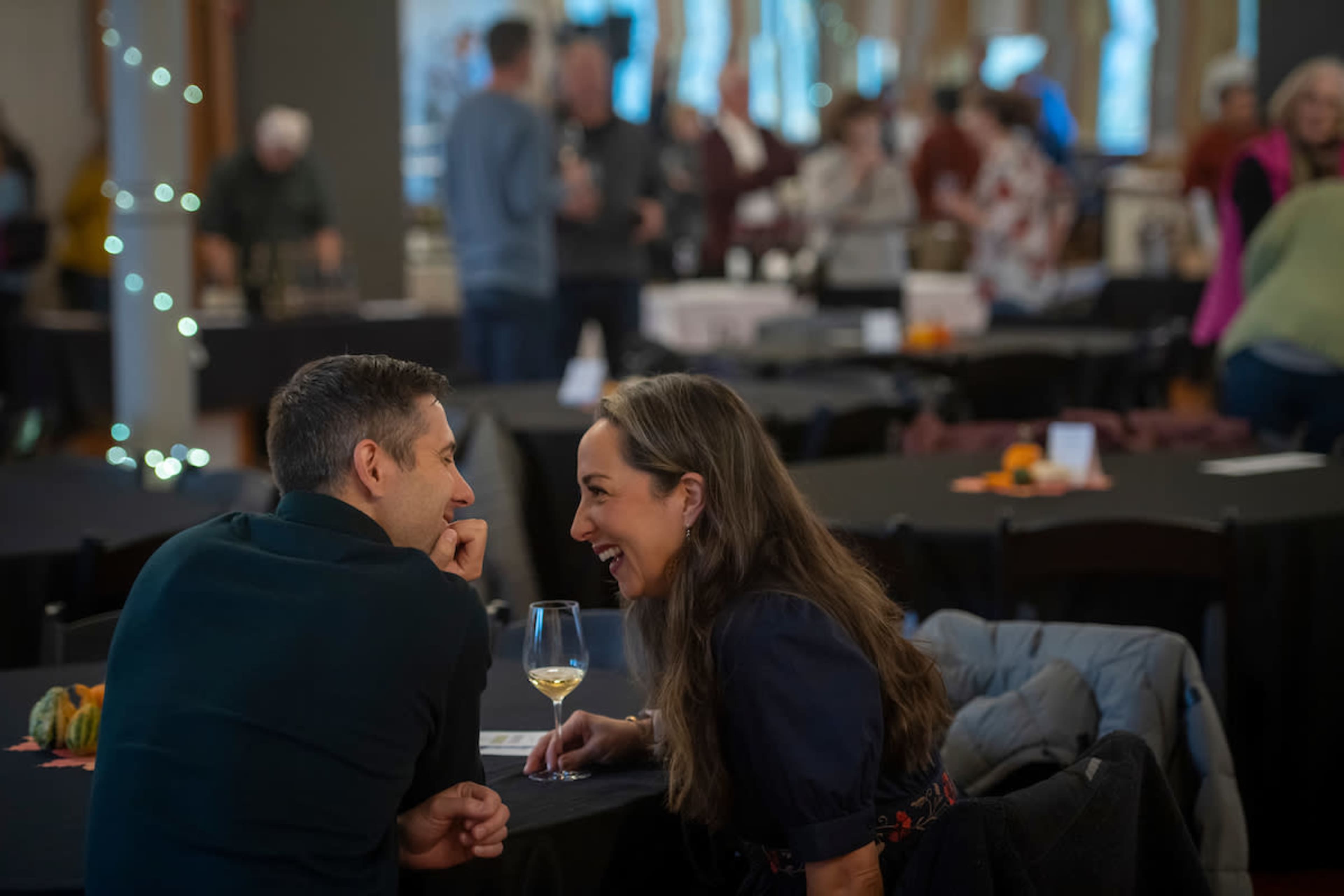 A couple sits close together at a table, laughing and enjoying each other's company in a crowded event space.