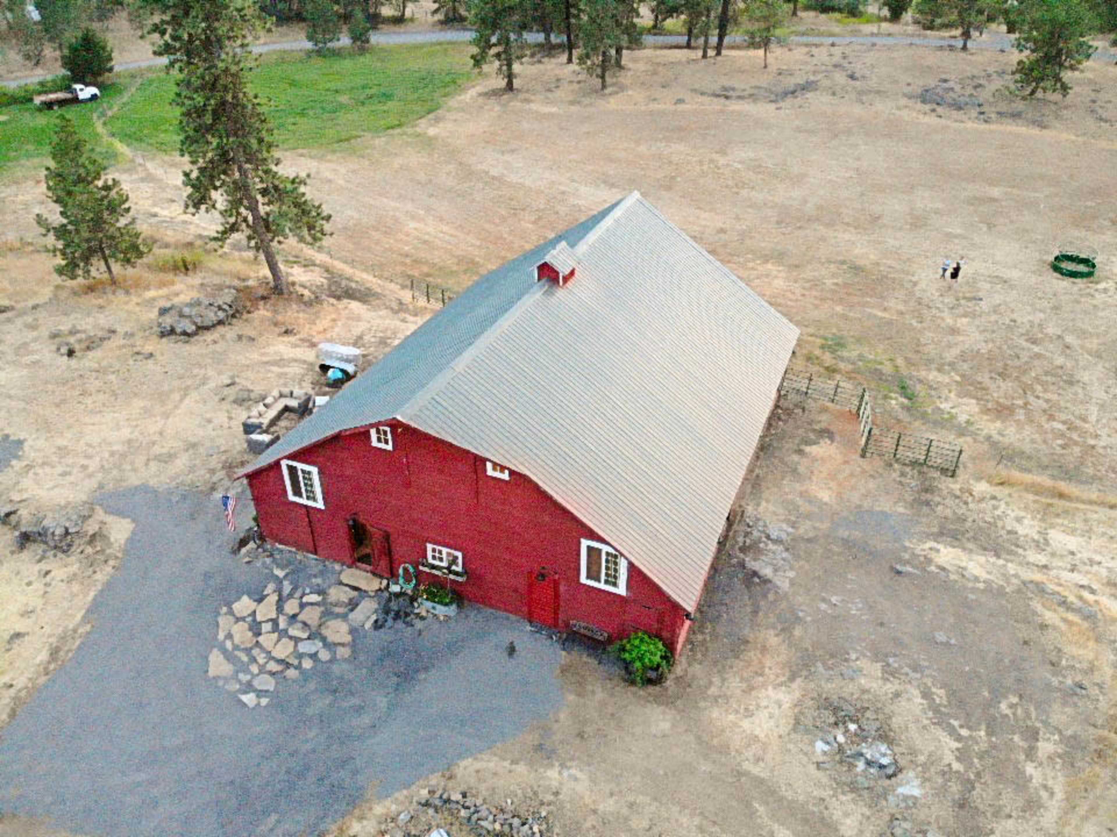 An aerial view shows a red barn surrounded by dry, open land with sparse vegetation and a gravel pathway leading to the entrance.