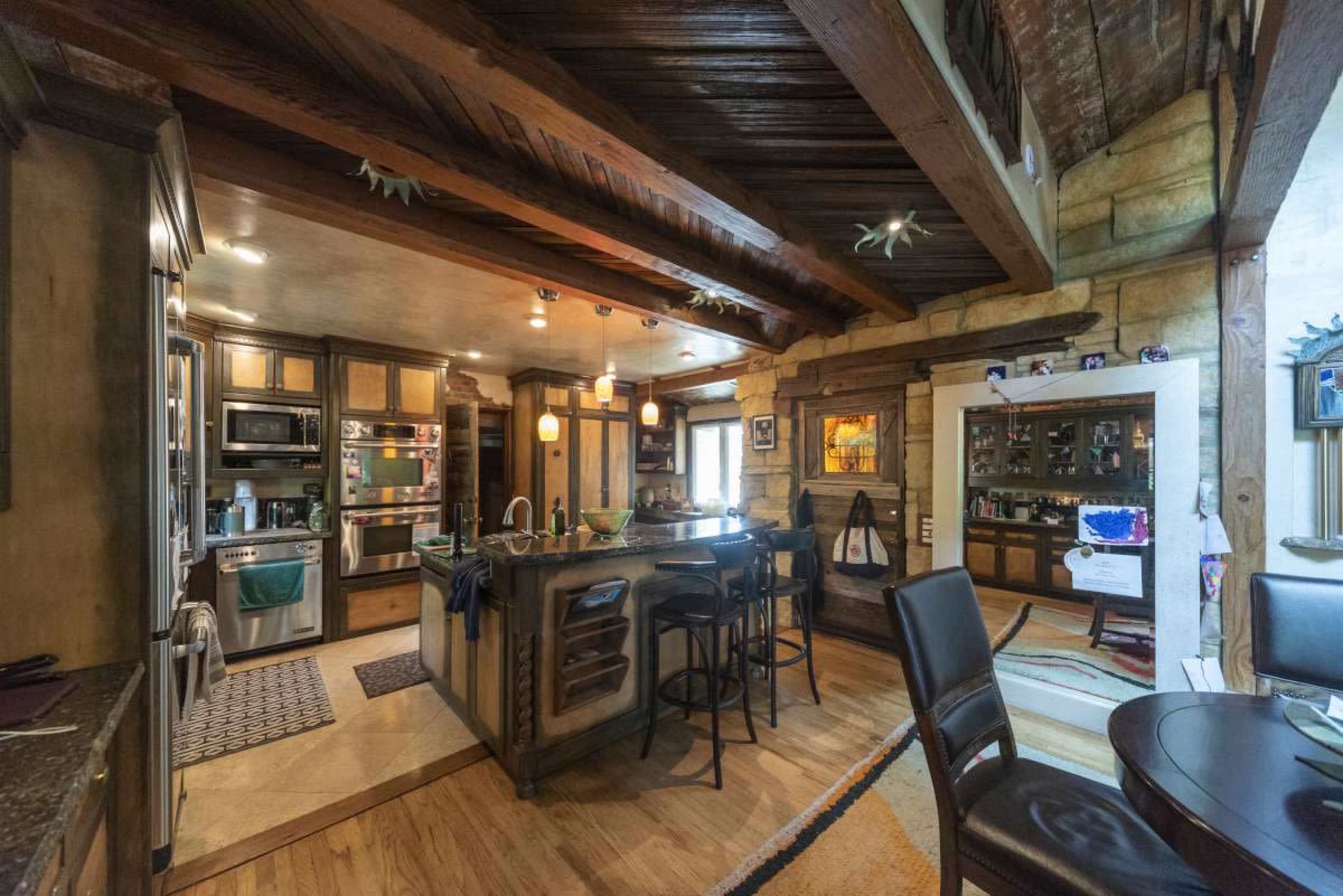 The image shows a rustic kitchen featuring wooden beams, an island with seating, and various appliances and cabinetry.