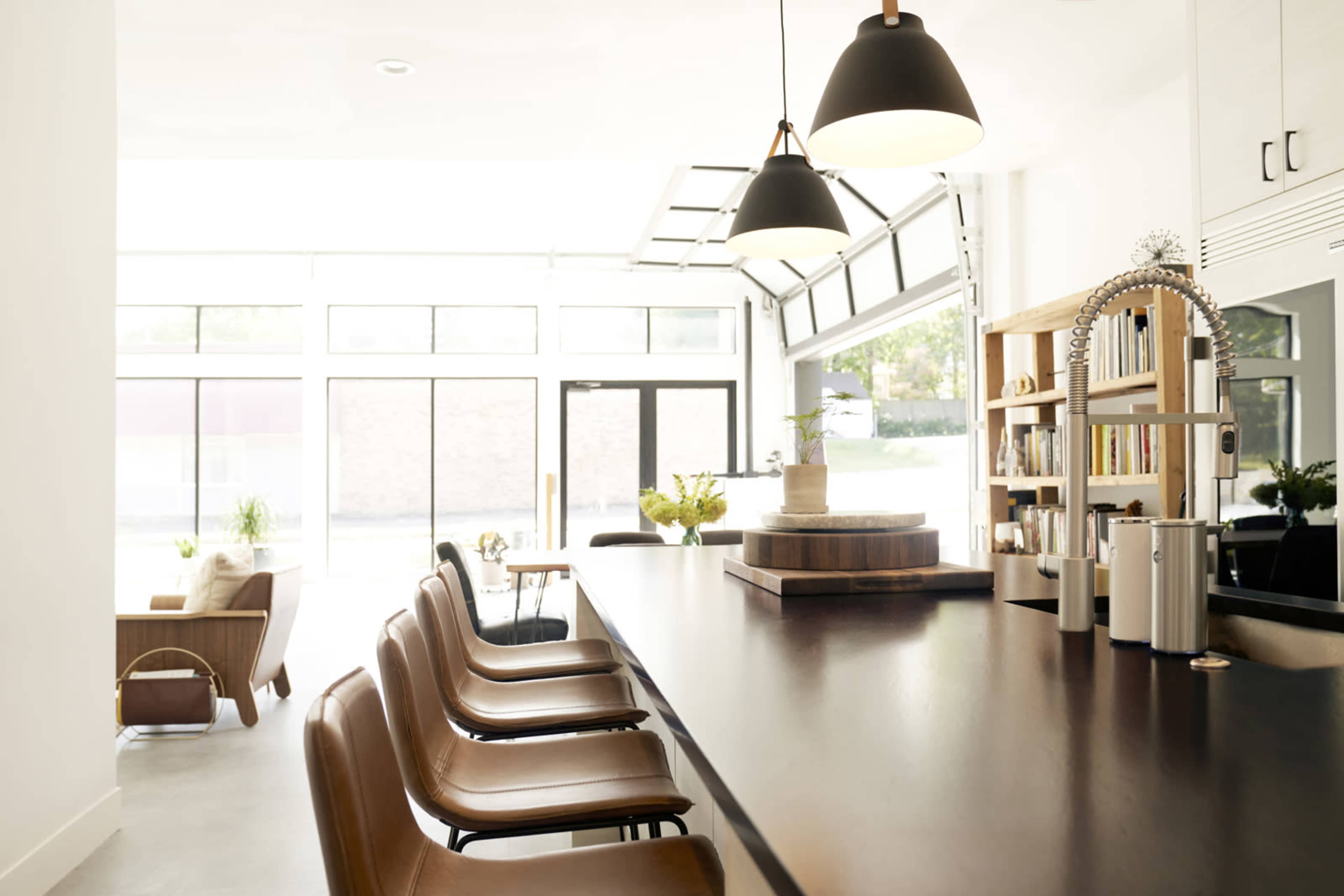 The image shows a modern café interior with a long wooden counter featuring several leather chairs, bright natural light streaming through large windows, and a seating area with a chair and table in the background.