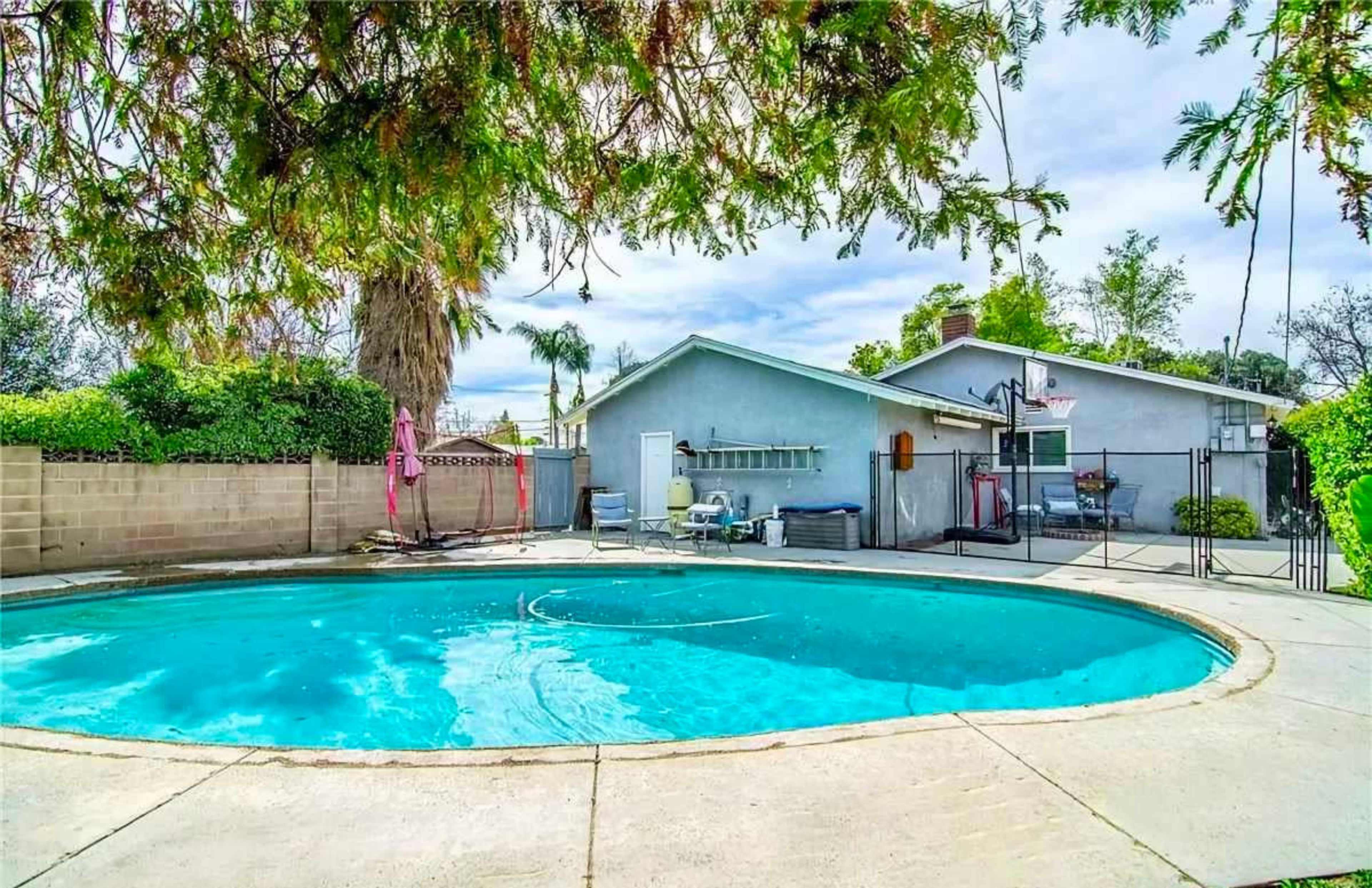 The image shows a pool surrounded by a concrete patio, with a residential house and foliage in the background.