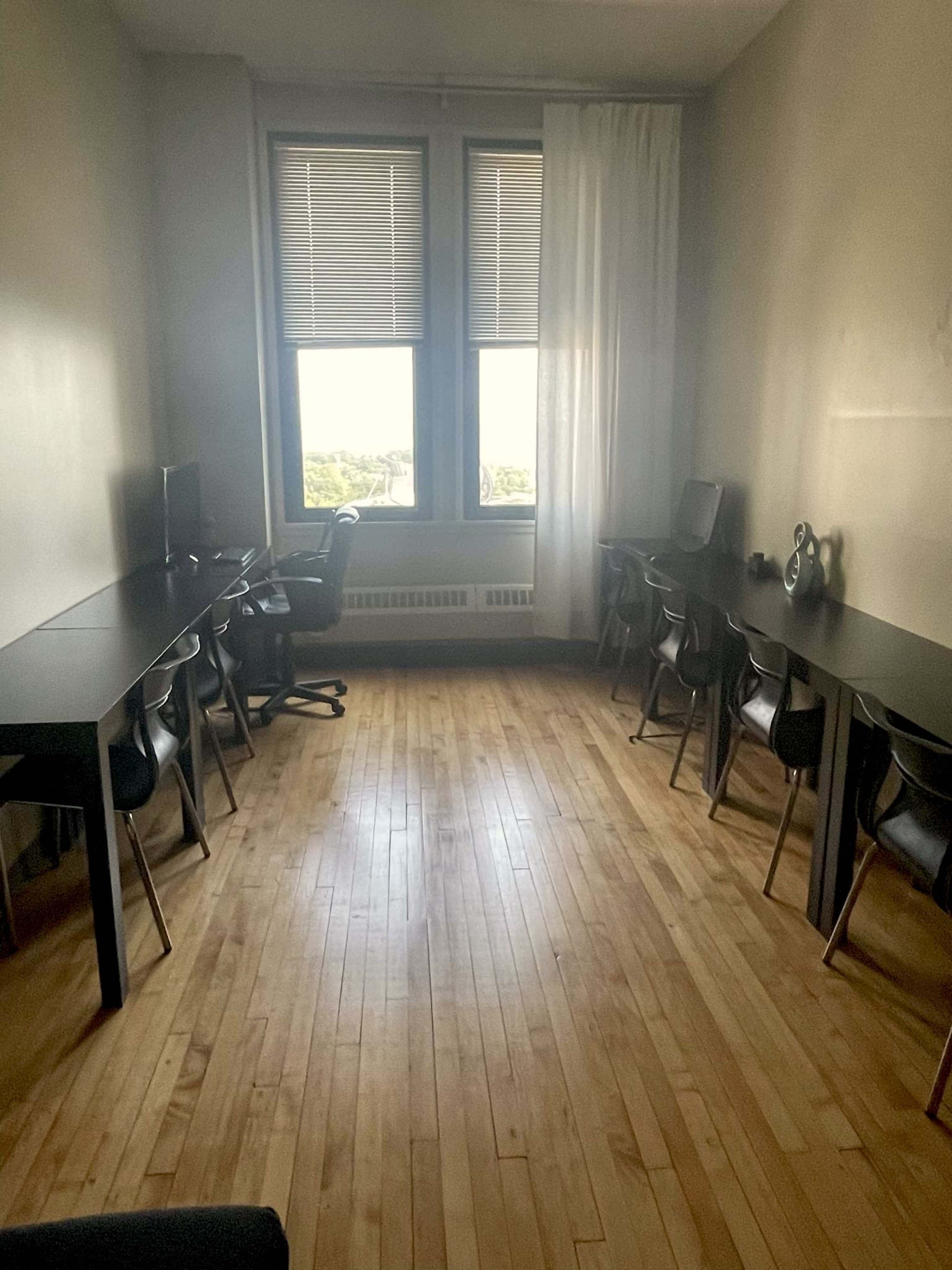 The image shows a spacious, minimalistic room with wooden flooring, containing two desks with chairs positioned along each side and large windows allowing natural light.