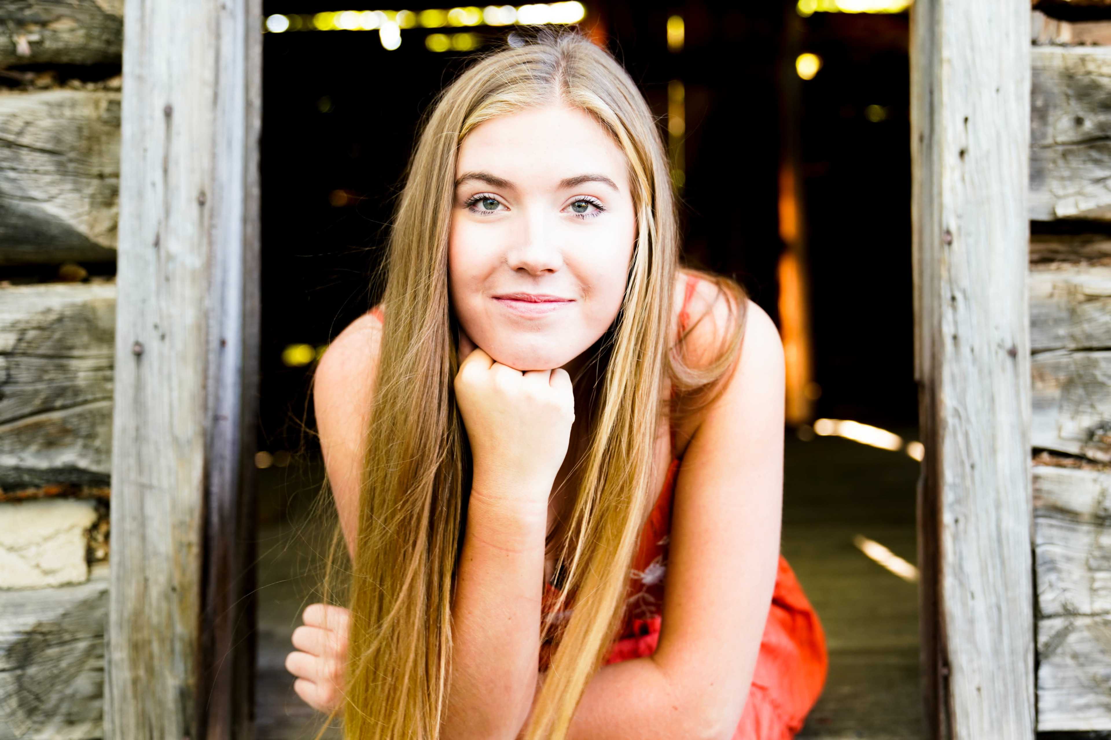 A young woman with long hair rests her chin on her hand while sitting in the doorway of a rustic wooden structure.