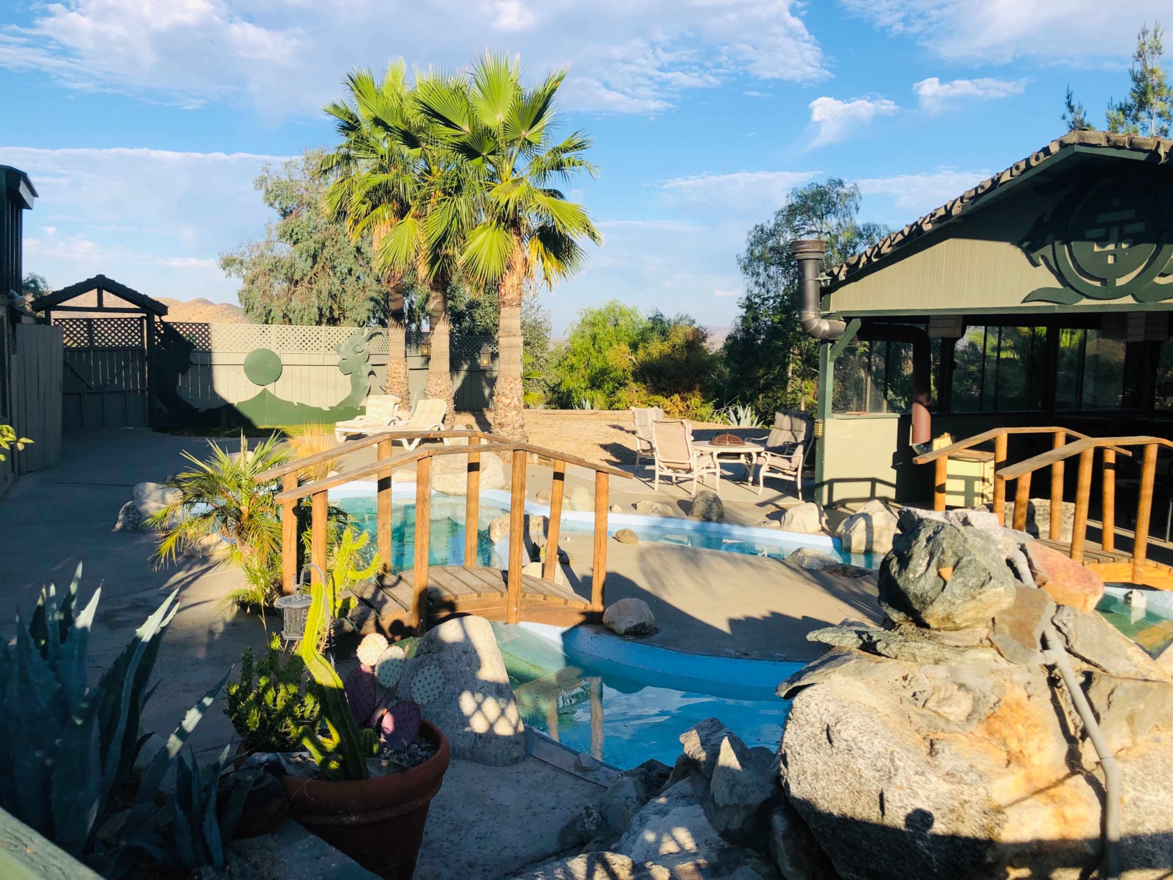 The scene shows a pool area with a wooden bridge, surrounded by palm trees and lounge chairs, along with a decorative rock feature.